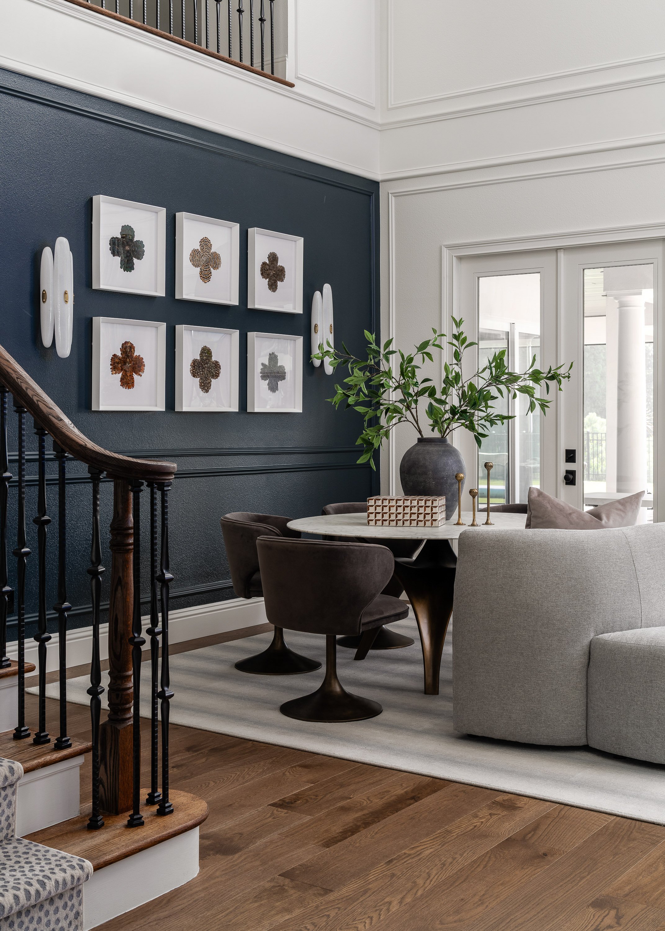 Inside view of a living room with a dark blue accent wall decorated with framed artwork of various textures and a white ceramic wall sculpture. A round table is surrounded by brown and ivory chairs, with a large gray sofa nearby. A black ceramic vase with green plant, gold candlesticks, and a decorative object are on the table. Natural light enters through glass doors.