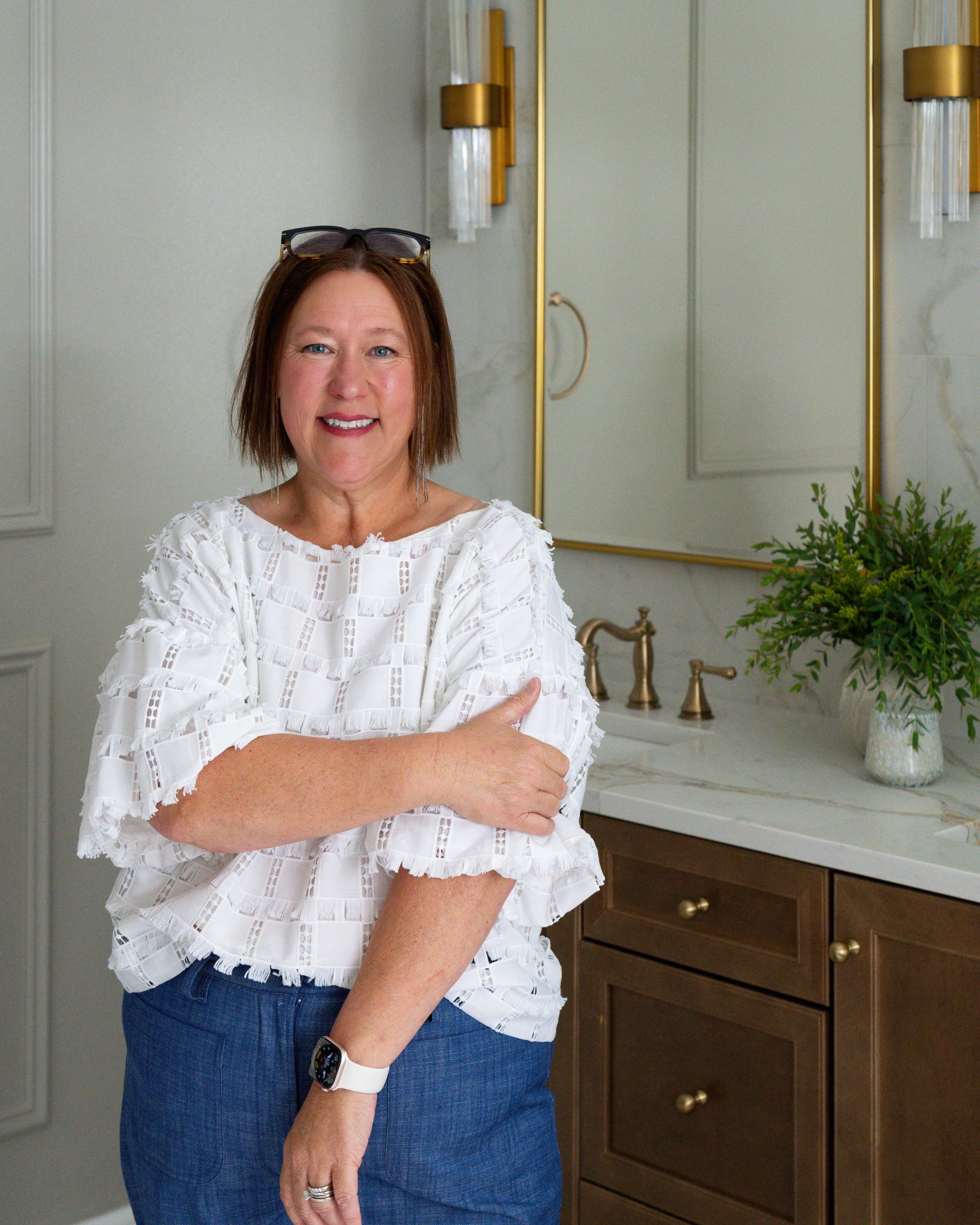 A woman with shoulder-length brown hair, smiling, wearing a white skirt, standing next to a warm wood bathroom vanity with gold fixtures and mirror.
