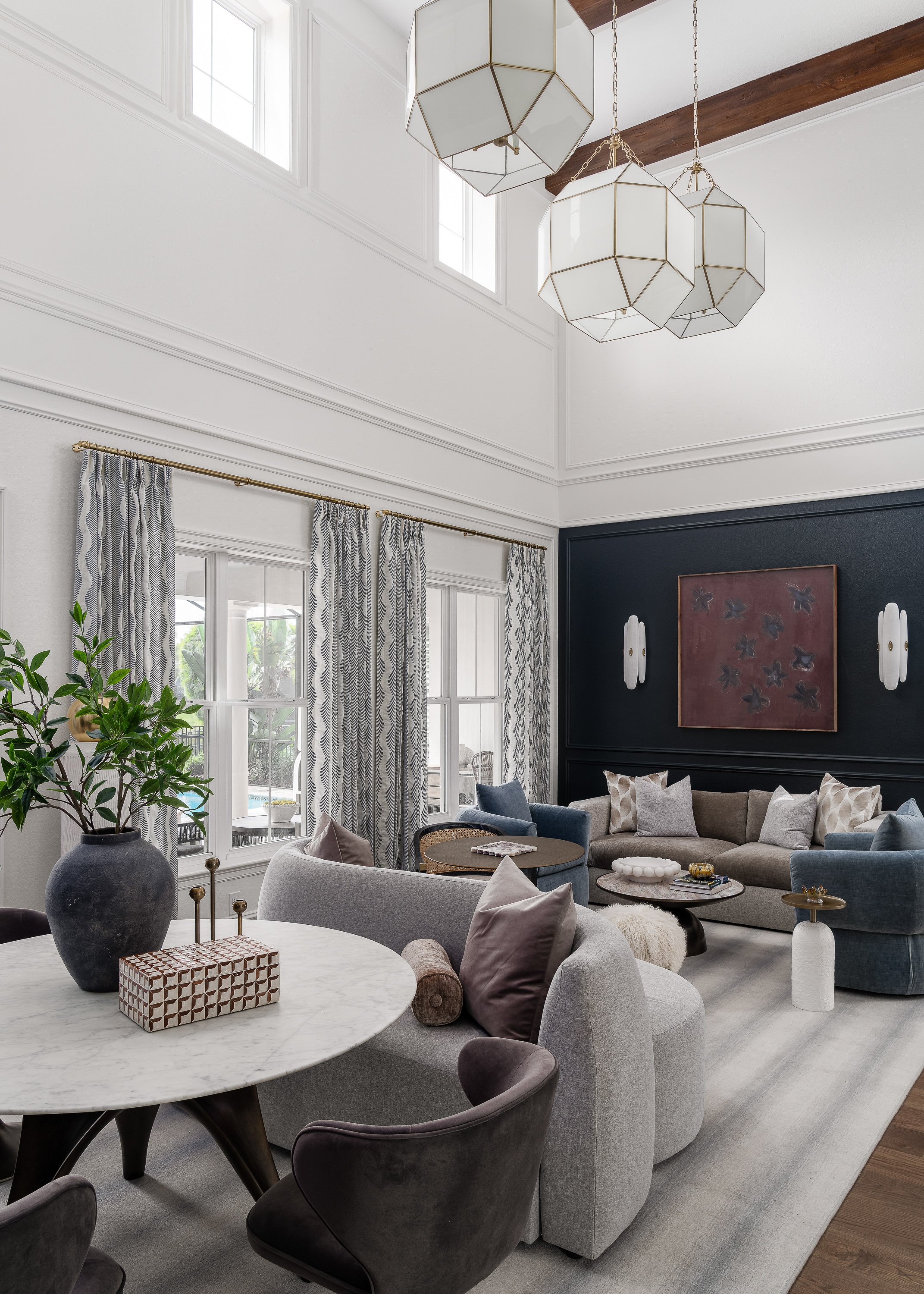 Modern living room with large windows, navy accent wall with artwork, beige sectional sofa with pillows, coffee tables, and a dining table with chairs. Decorative items include a large plant in a dark vase, candles, and decorative objects. The ceiling features a geometric pendant light, and there is wood ceiling beam and white walls.