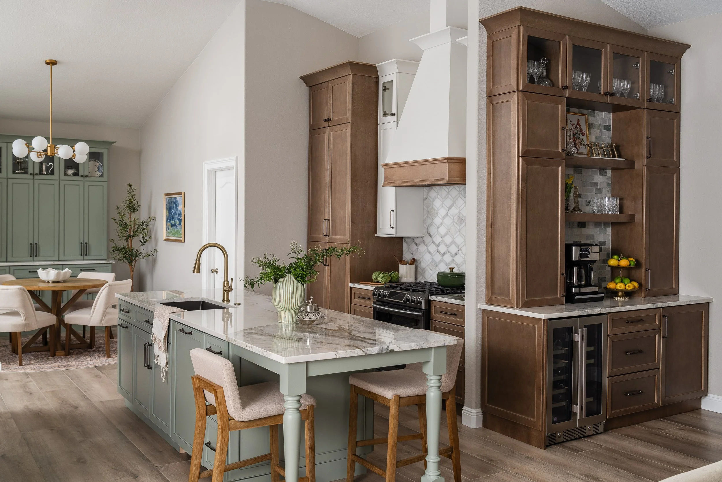 A modern kitchen with a large island featuring a marble countertop, light blue cabinetry, and a brass faucet. To the right, brown wooden cabinets house glassware and a wine cooler. In the background, a dining area with a round wooden table and beige chairs is visible, along with light green cabinets and a chandelier.