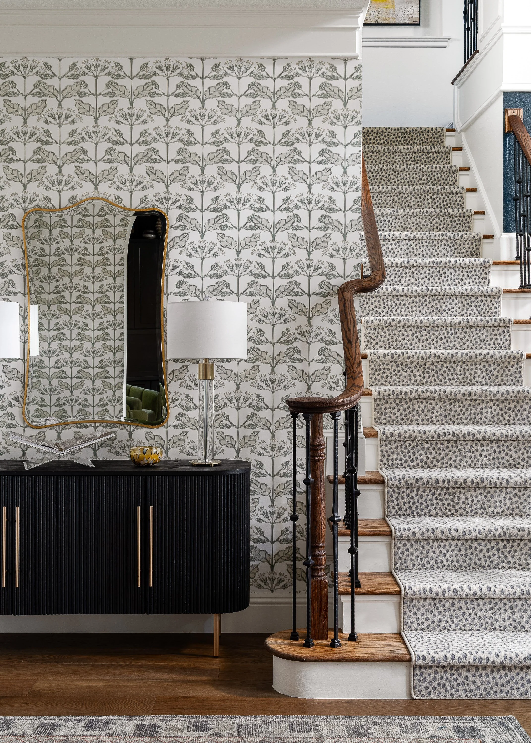Interior of a house showing a staircase with a patterned carpet runner, a black sideboard with decorative objects, a mirror, a lamp, and a wall with floral wallpaper.