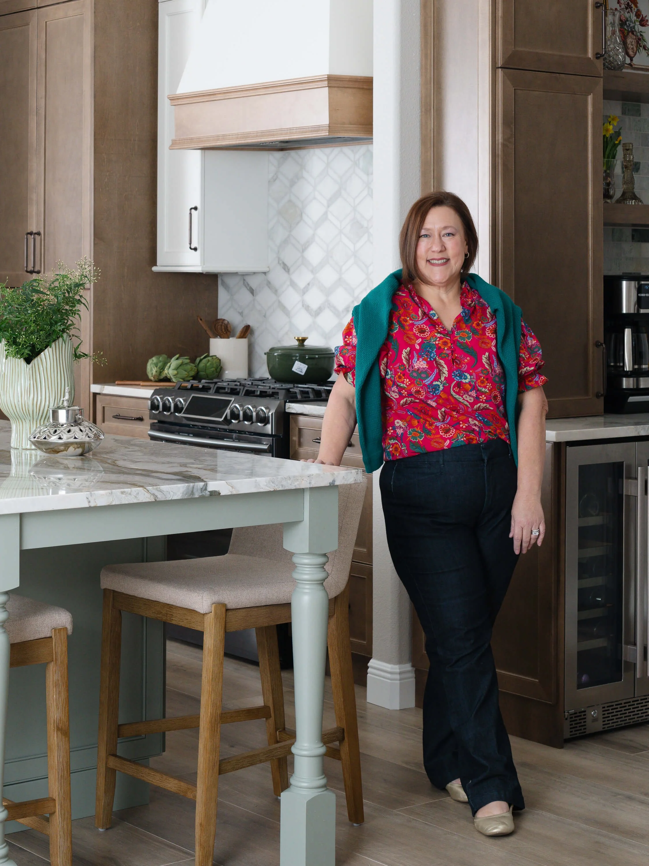 A woman standing and leaning against a kitchen island in a modern kitchen with wooden cabinets, a white marble countertop, and decorative items on the shelves.