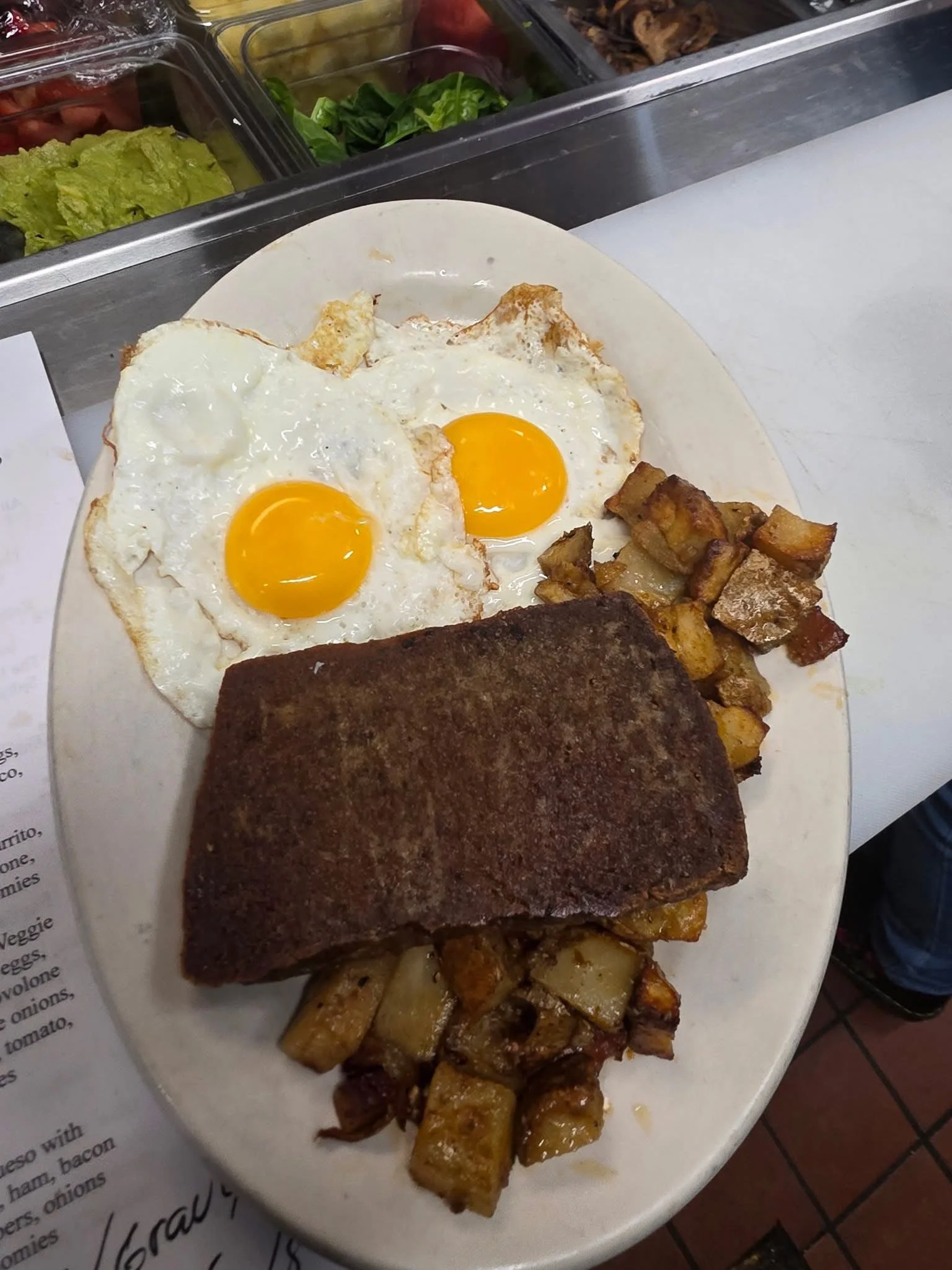 A plate of breakfast food with two sunny side up eggs, roasted potatoes, a piece of corned beef, and crispy bacon pieces, on a white plate.