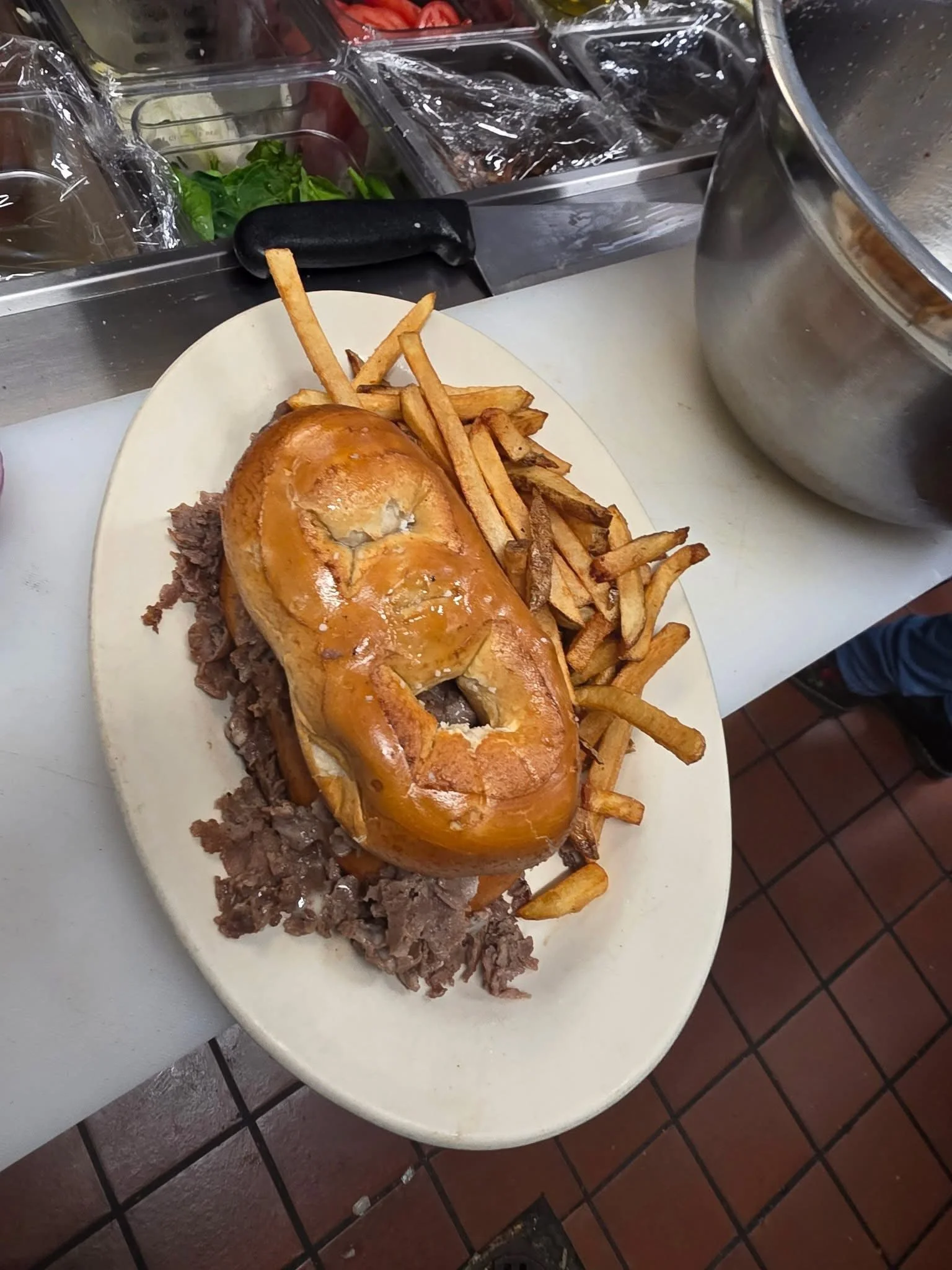 A plate of food with chopped roast beef, french fries, and a pretzel bun filled with roast beef, on a white tray in a restaurant or diner setting.