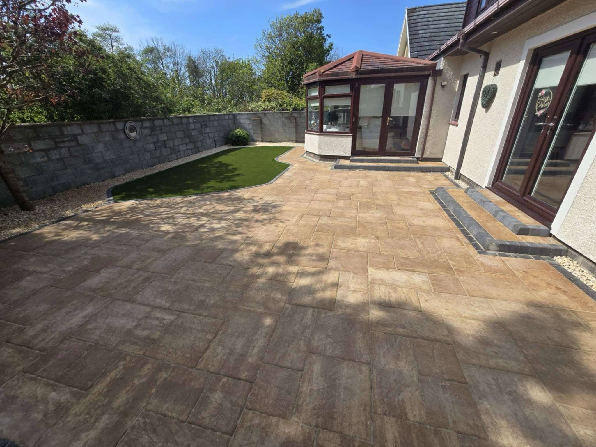 A backyard patio with stone tiles, a small artificial grass patch, a brick wall in the background, and a sunlit house with a conservatory and sliding glass doors.