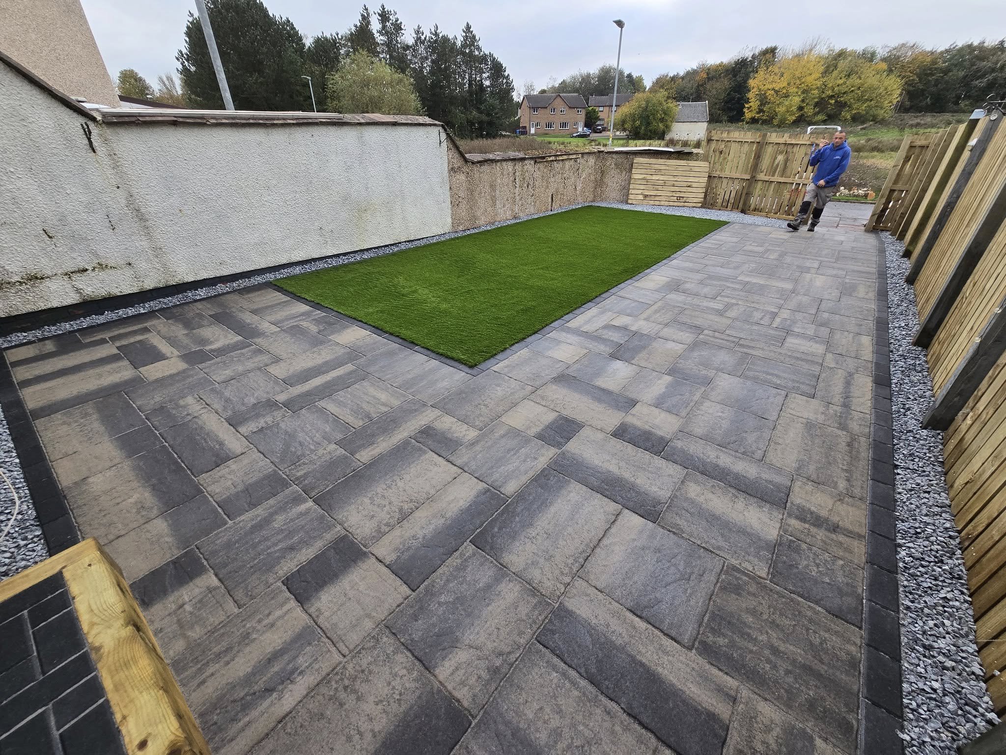 backyard with stone patio, a rectangular patch of artificial grass, a wooden fence, and a person standing outside near the gate.