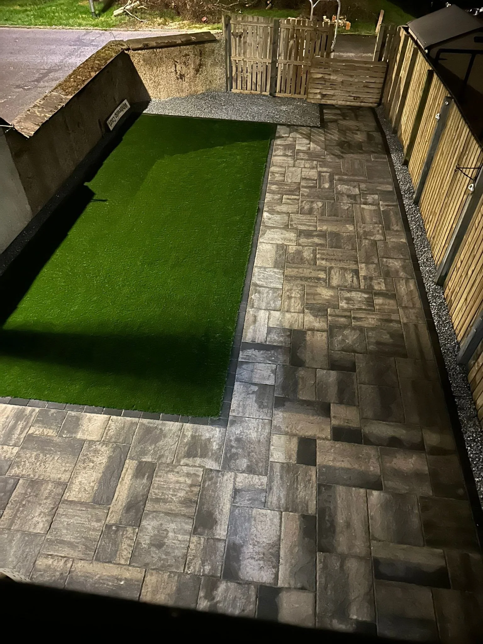 A small backyard patio at night with a section of artificial grass, stone pavers, a wooden fence, and a wooden gate.