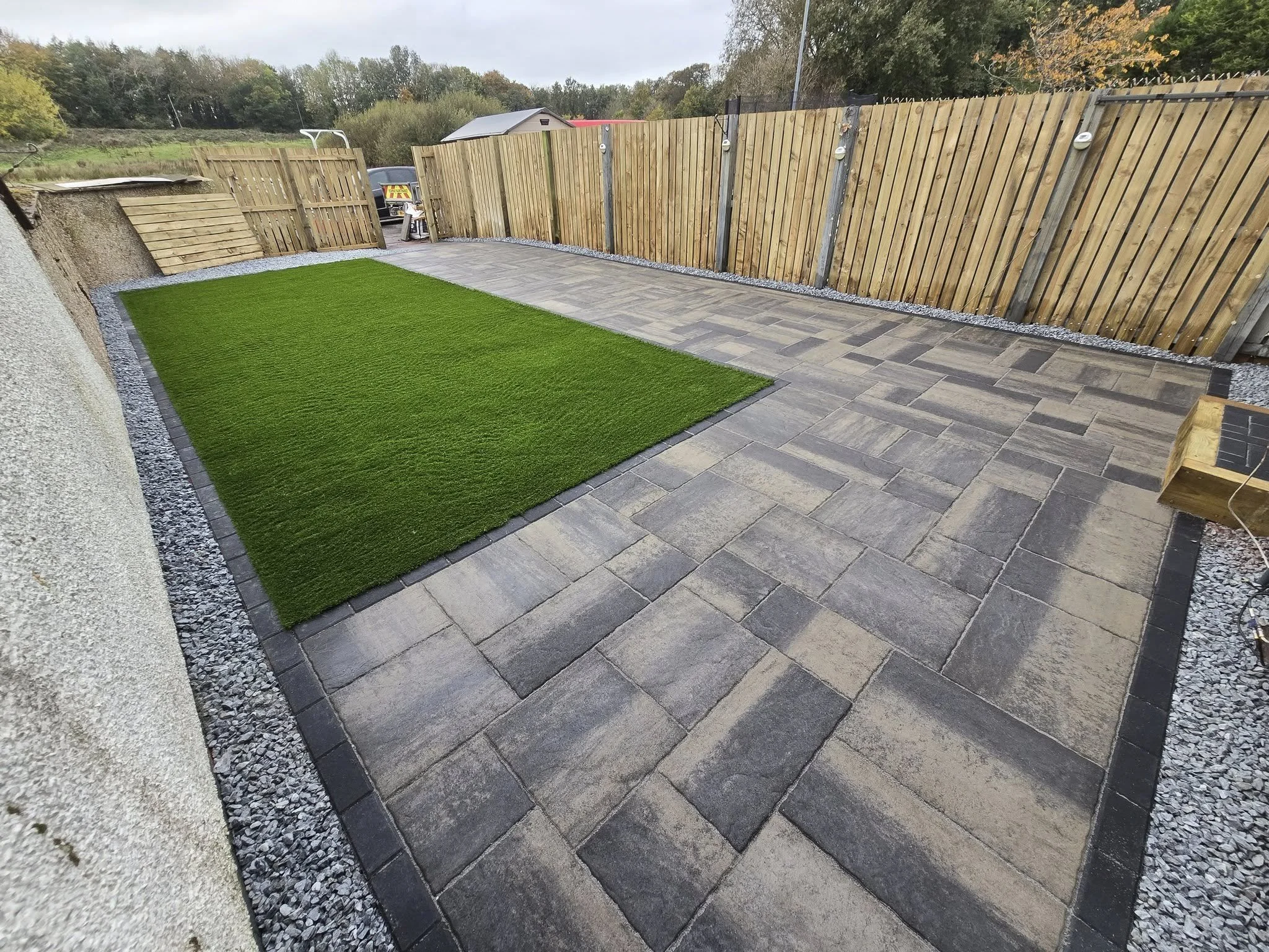 Backyard patio with a grassy patch and stone paving, enclosed by a wooden fence, with trees and a shed in the background.
