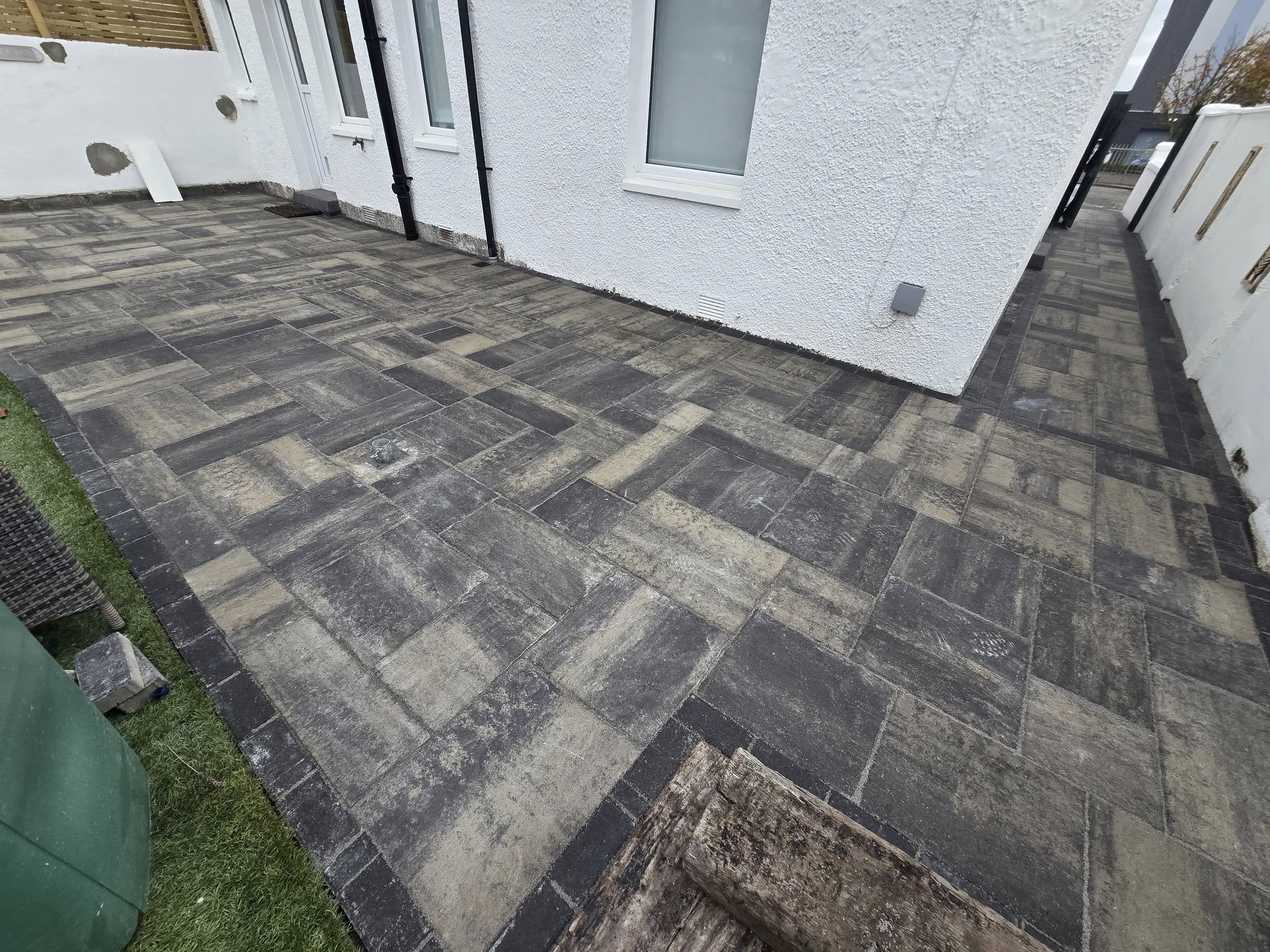 Newly paved outdoor patio area with dark rectangular stone tiles surrounding a white house, featuring a sliding door, windows, and some construction materials and tools nearby.
