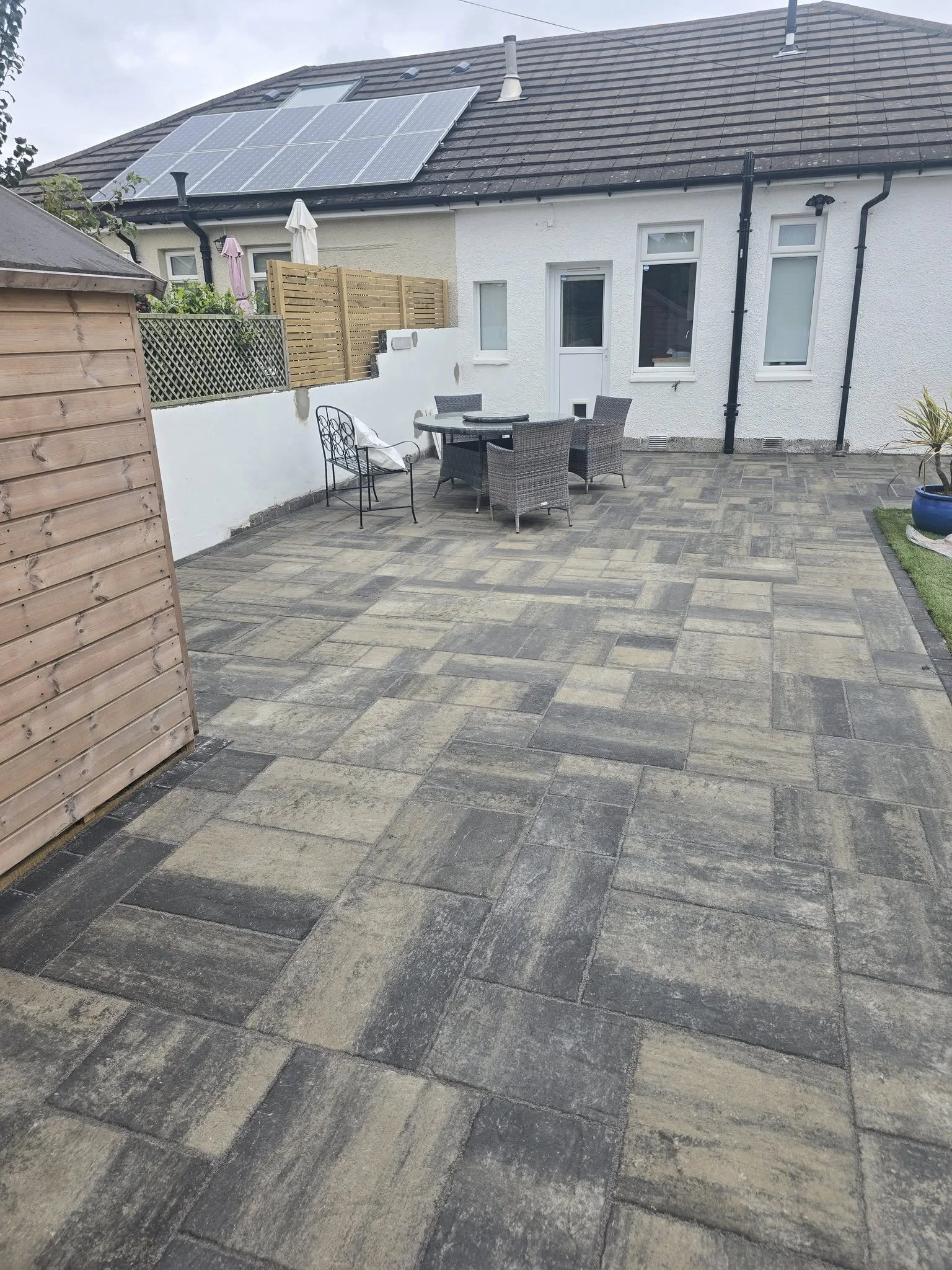 Backyard patio with stone paving, outdoor dining table with four chairs, and a wooden shed. The house has solar panels on the roof and a white exterior wall with a door and windows.