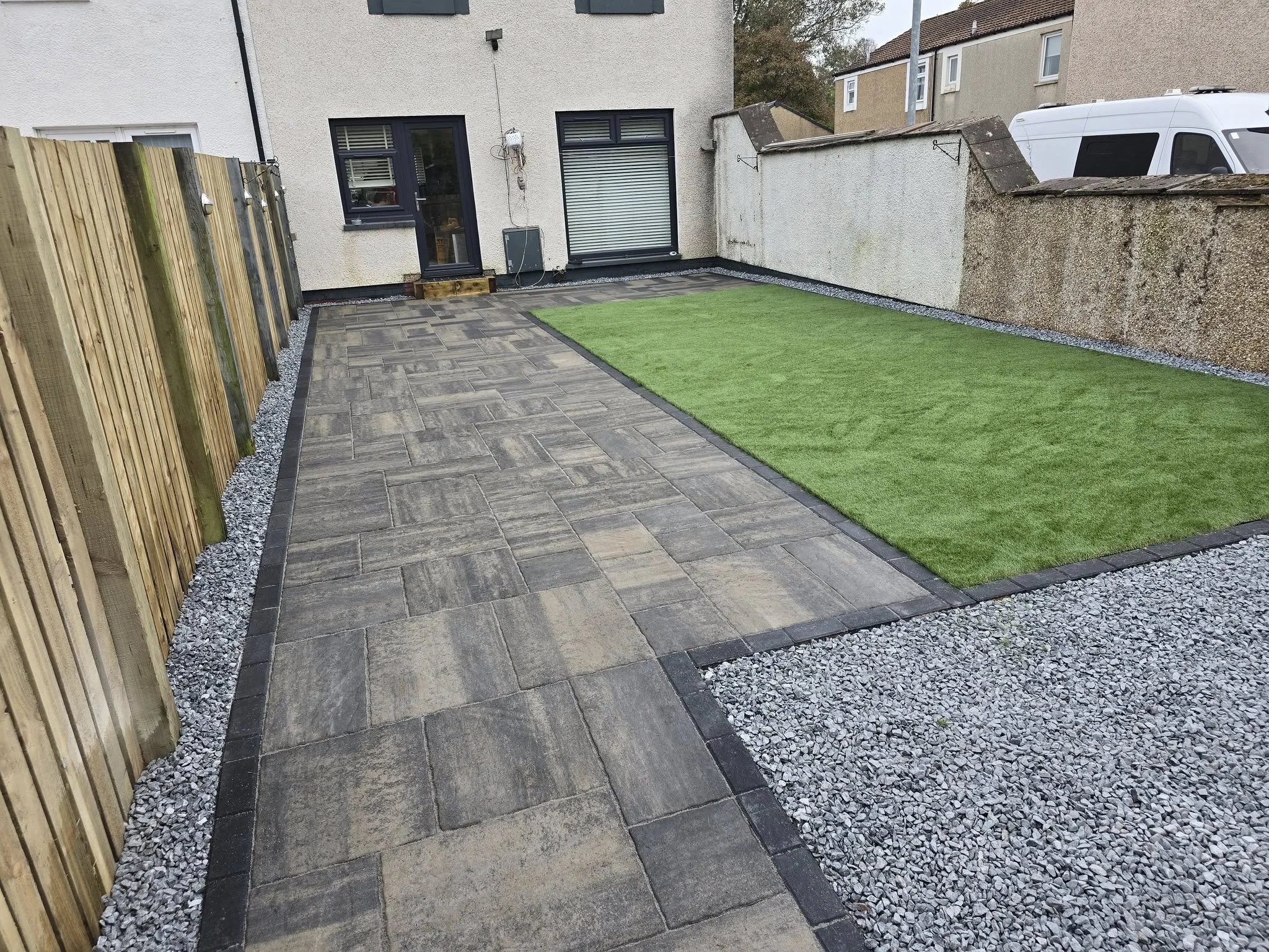 Backyard with paved walkway, artificial grass, gravel borders, wooden fence on one side, concrete wall on the other, and a beige house with black window frames and a door in the background.