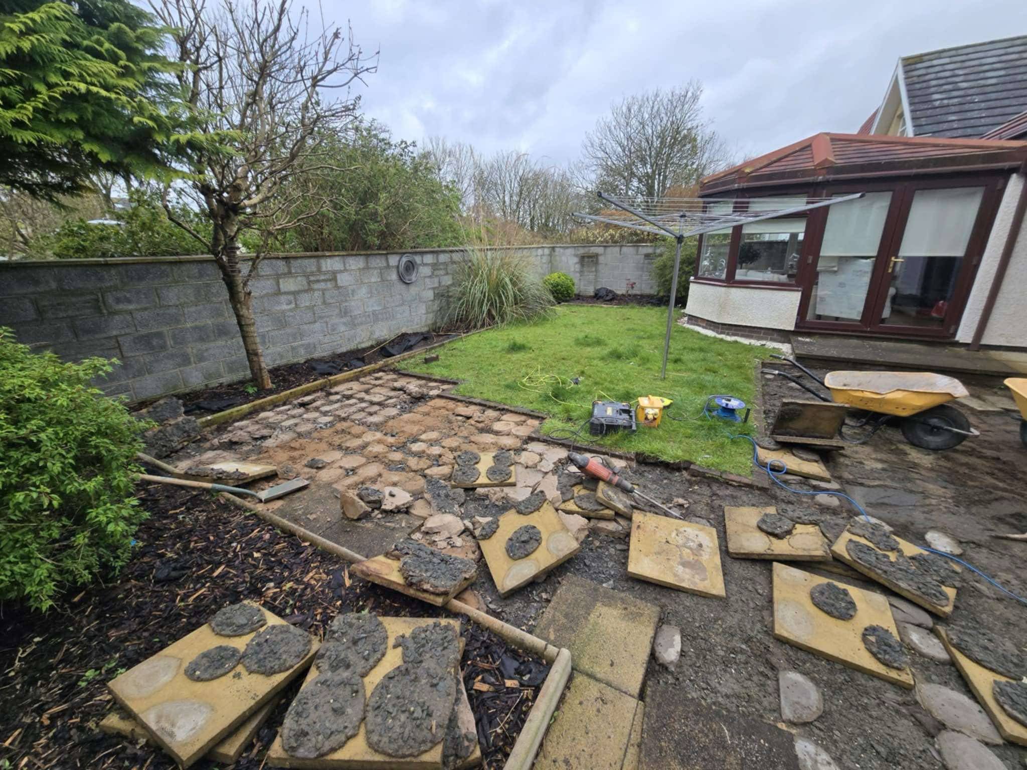 A backyard undergoing patio renovation with uneven paving stones, tools, and construction materials scattered around.