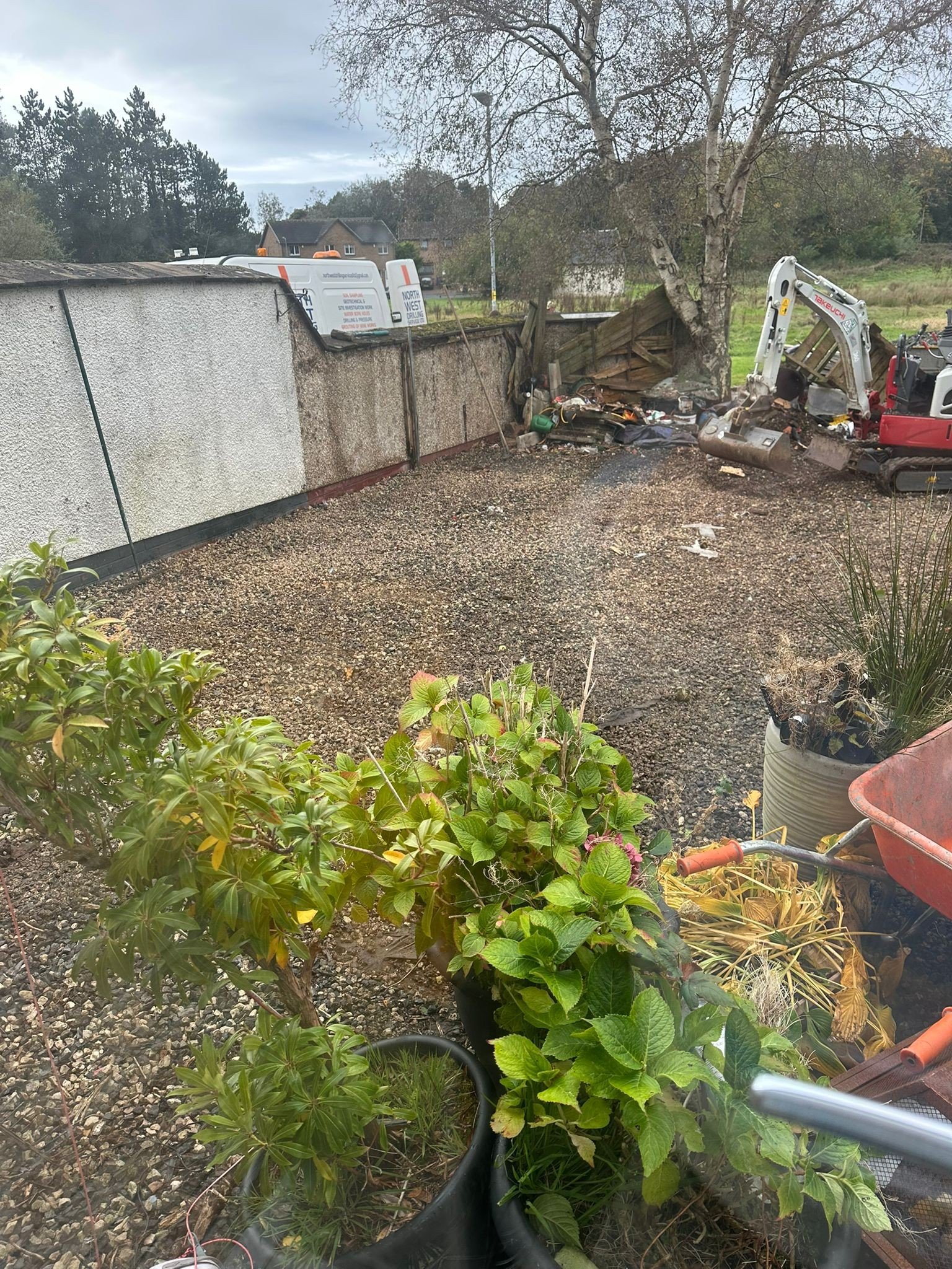 View of a backyard with a gravel surface, potted plants in the foreground, and a damaged fence with construction equipment and debris near it in the background.