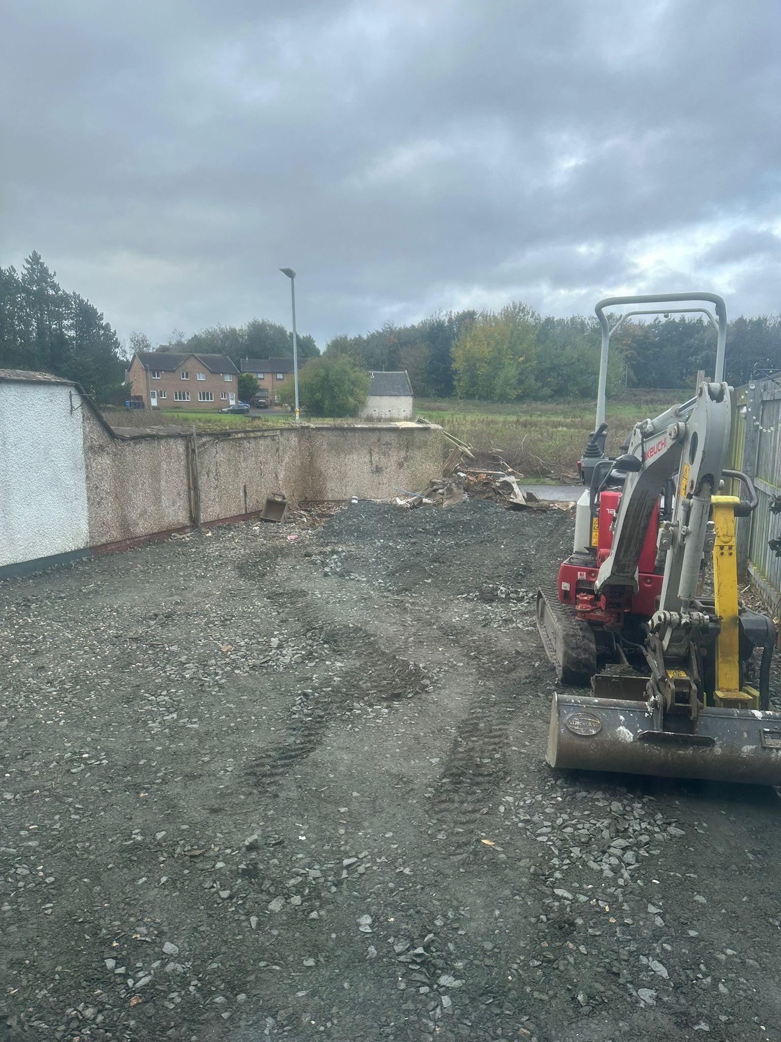 A construction site with a mini excavator on dirt ground and a concrete wall in the background, under cloudy sky.