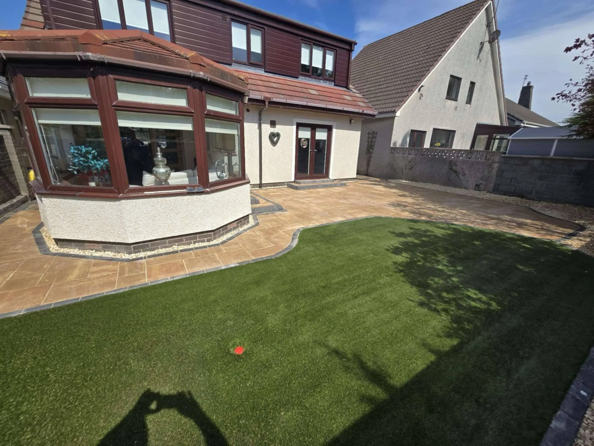 A backyard with a small lawn, stone patio, and a house with a bay window and glass door.