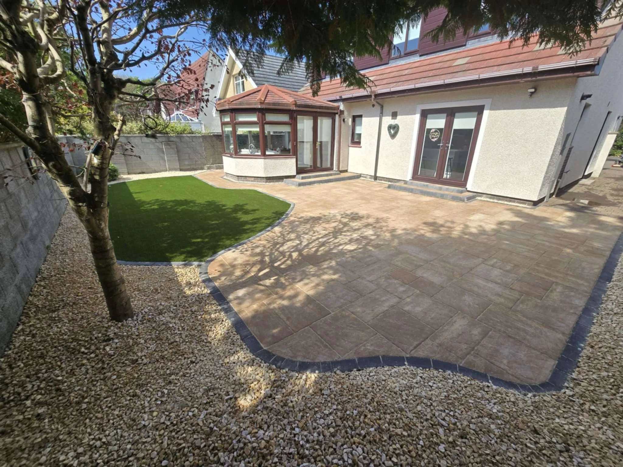 Backyard with a tree, artificial grass, a paved patio area, and a house with a glass porch and double doors.