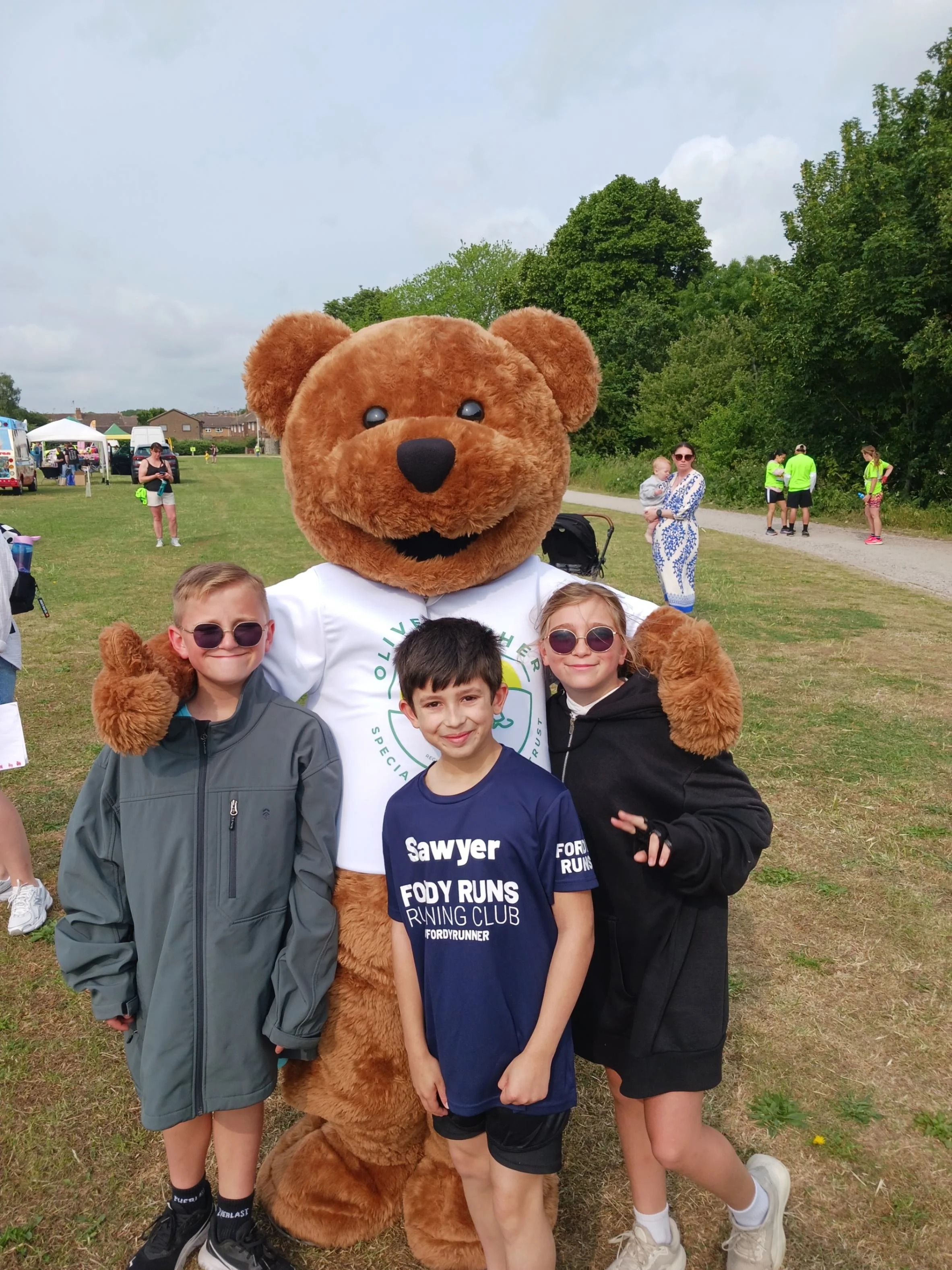 Children smiling and standing with the charity bear mascot at an outdoor community event