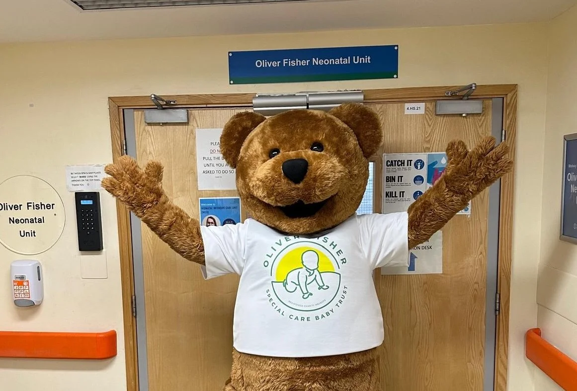 Oliver Fisher bear mascot costume standing with arms raised in front of hospital neonatal unit entrance