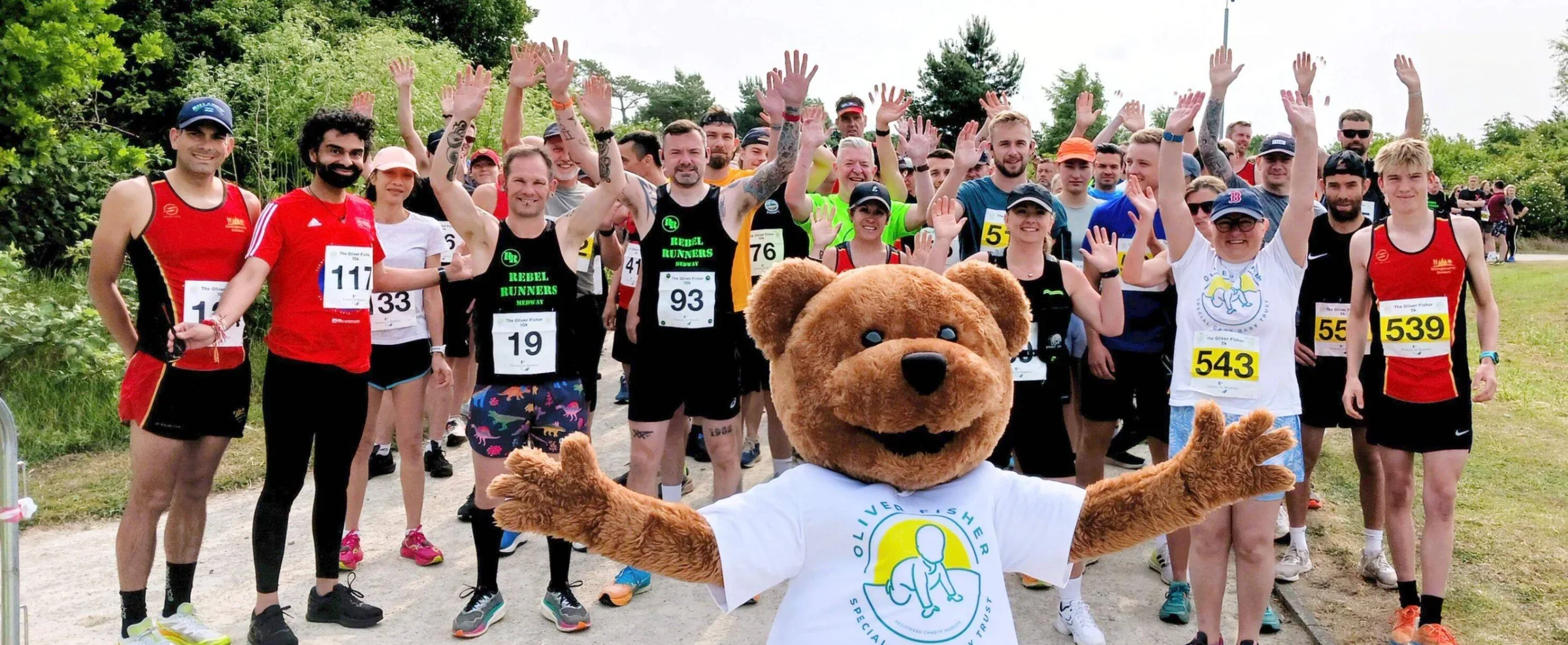 Group of runners at a charity event with the Oliver Fisher bear mascot waving among participants wearing race numbers