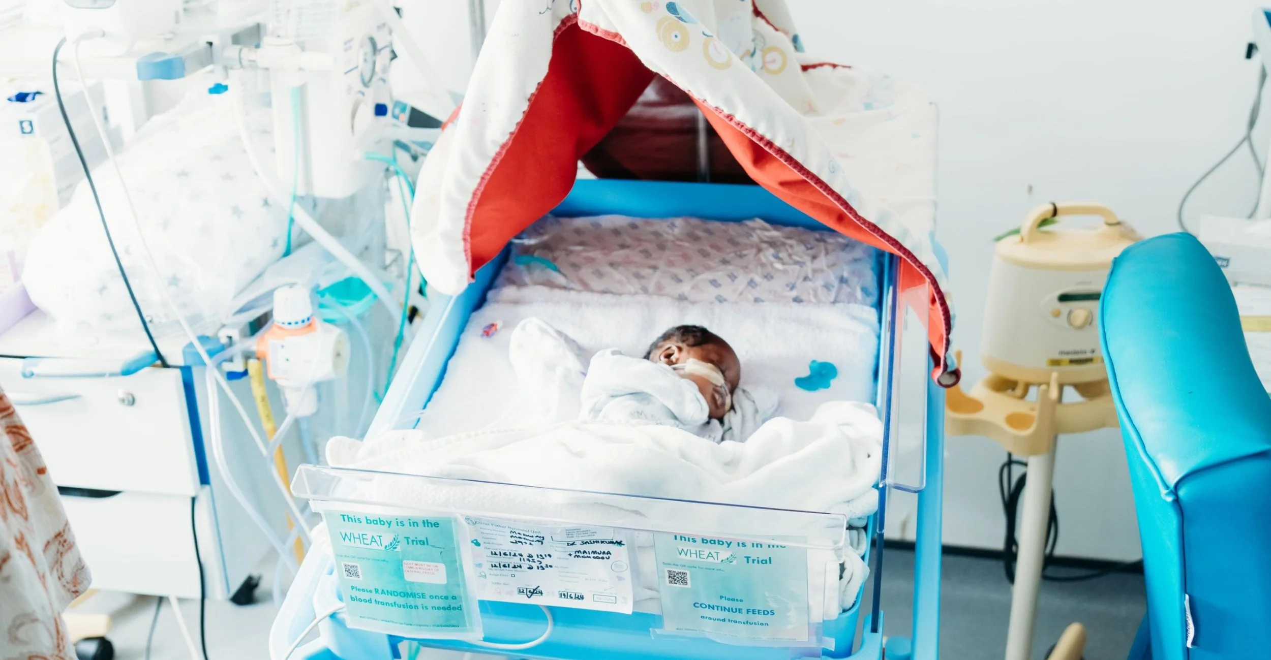 Baby resting in neonatal hospital cot surrounded by medical equipment and blankets
