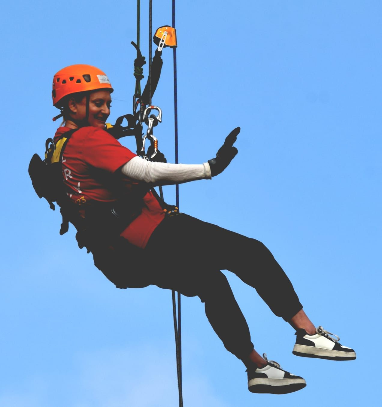 Photo of a women in a red tshirt, and orange hard hat, attached to a rope with a harness, doing an abseil