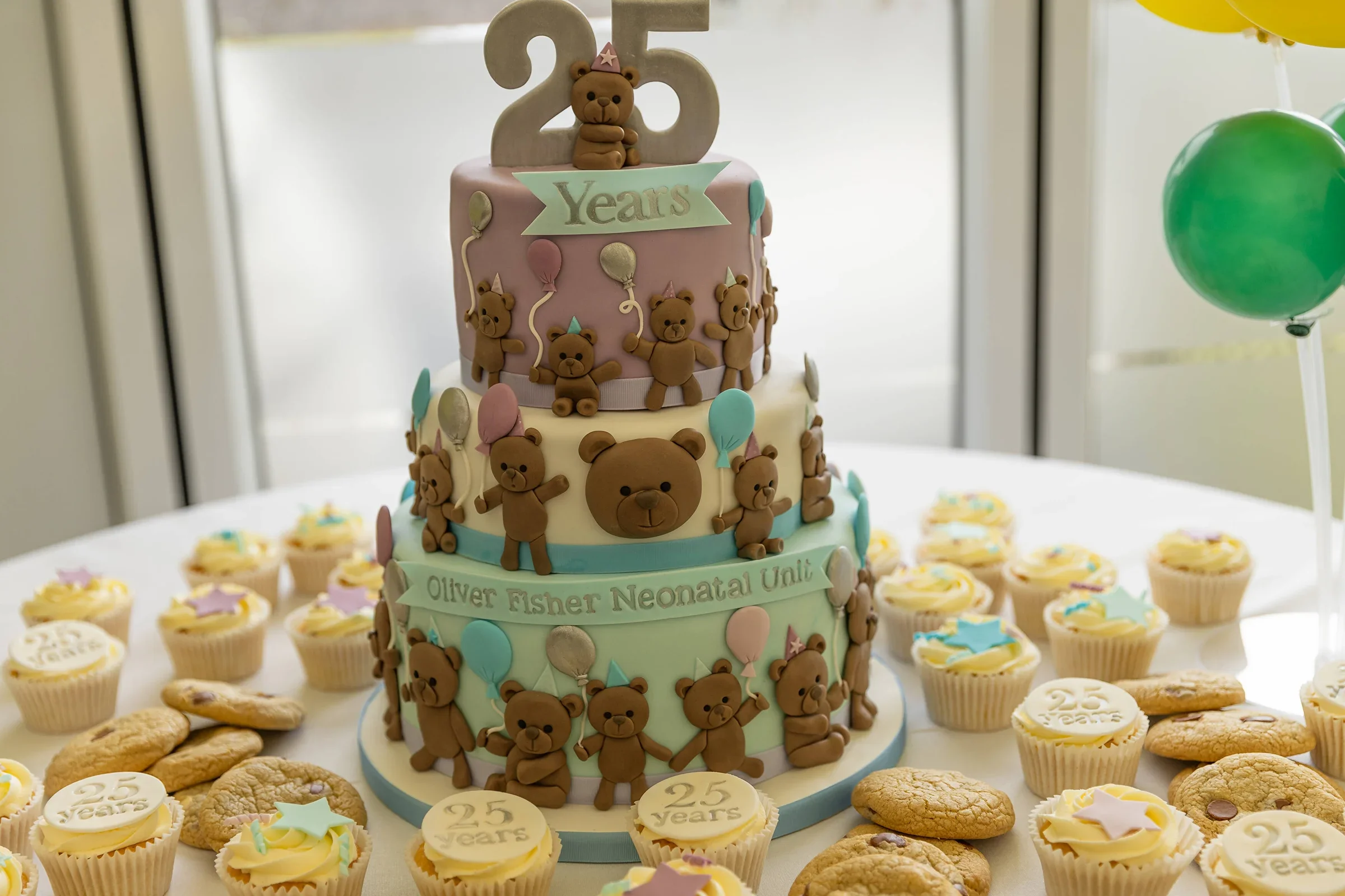 A photo of a 3 tier cake with 25 years and fondant bears all over. The cake is surrounded by cupcakes.