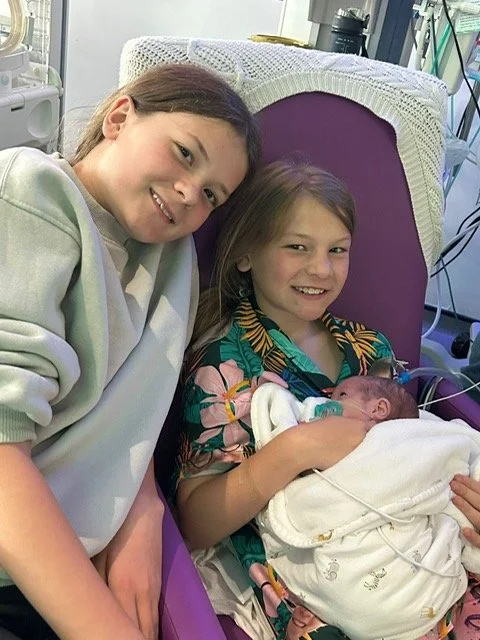 Two girls smiling on a hospital chair holding their baby sister who is in a blanket and connected to wires.