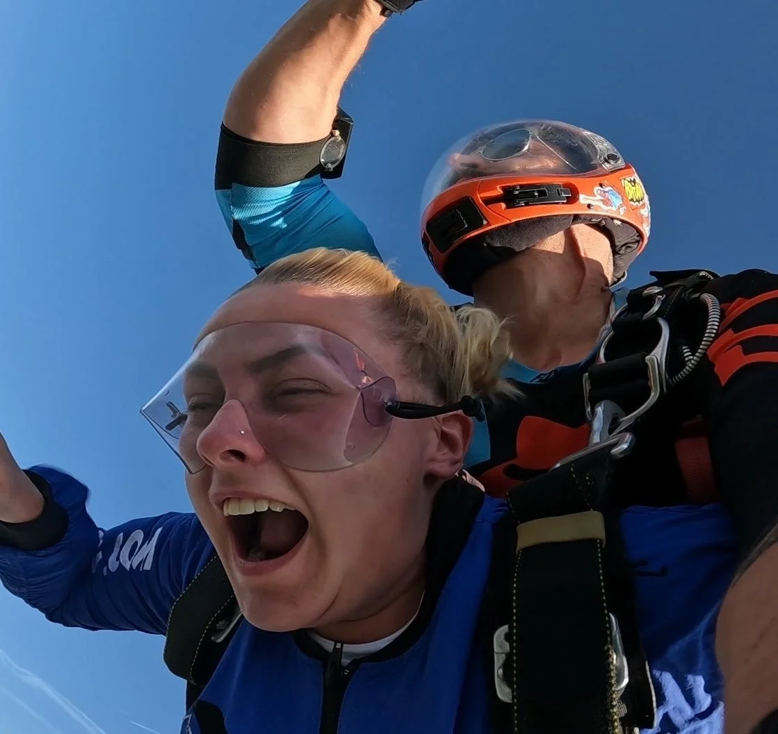 Woman mid skydive with an instructor wearing goggles and harness with clear blue sky in the background