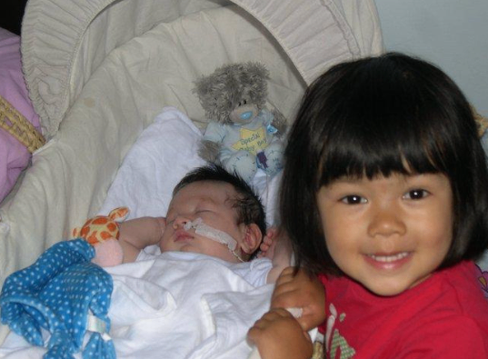 Alister asleep in a baby basket his sister stood next to him smiling