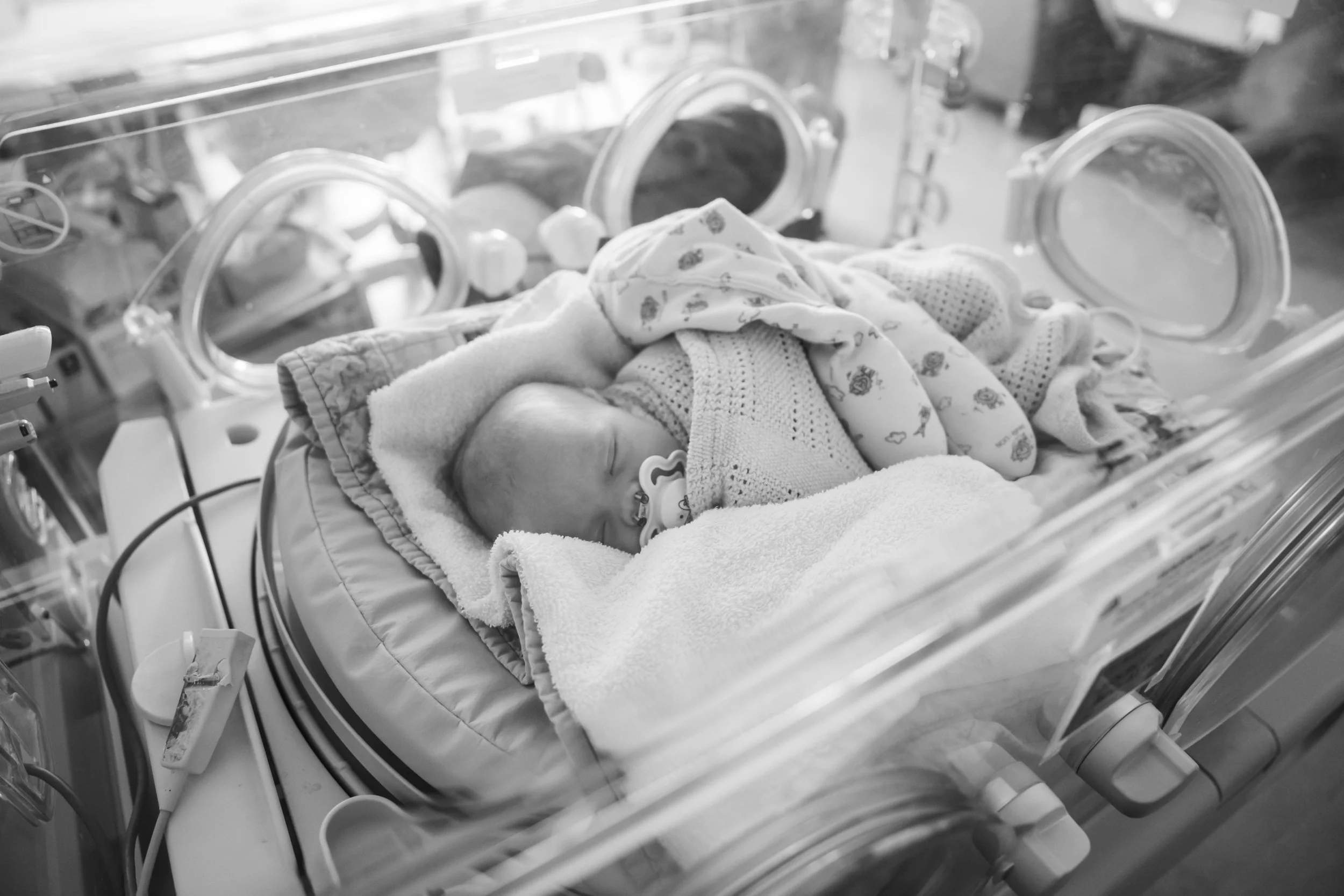 Sleeping newborn baby wrapped in blankets inside hospital cot shown in black and white