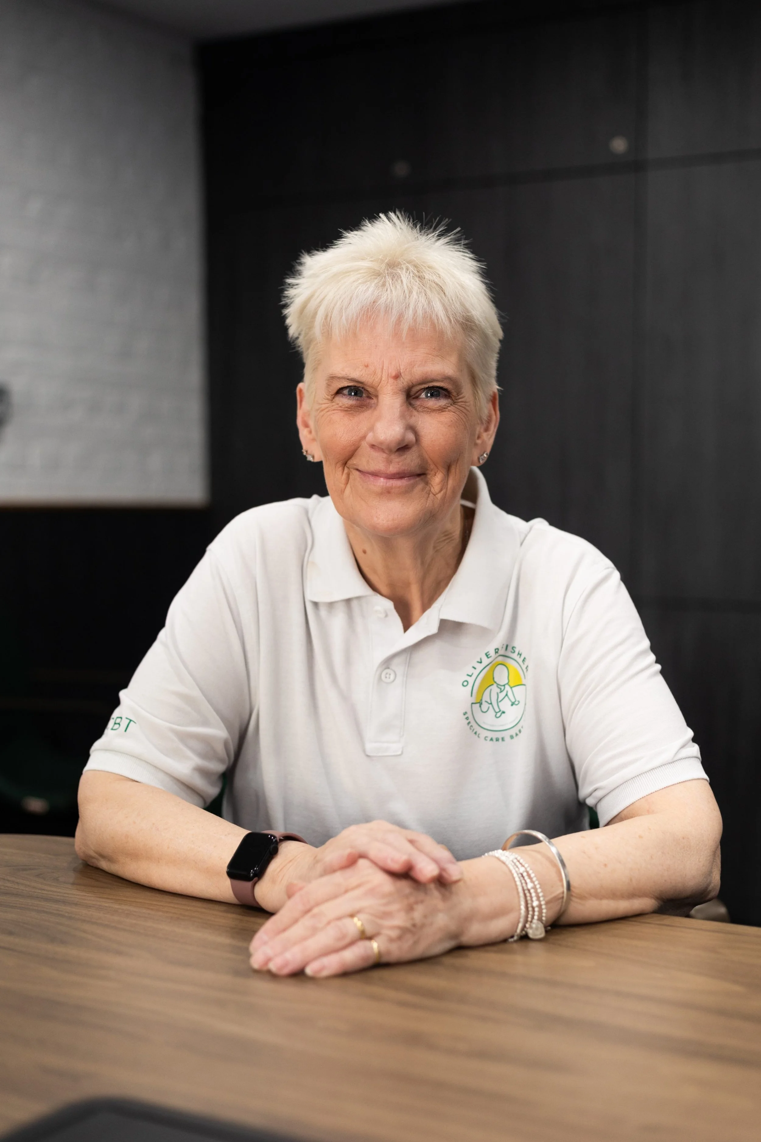 Woman smiling wearing white polo shirt with charity logo on plain background