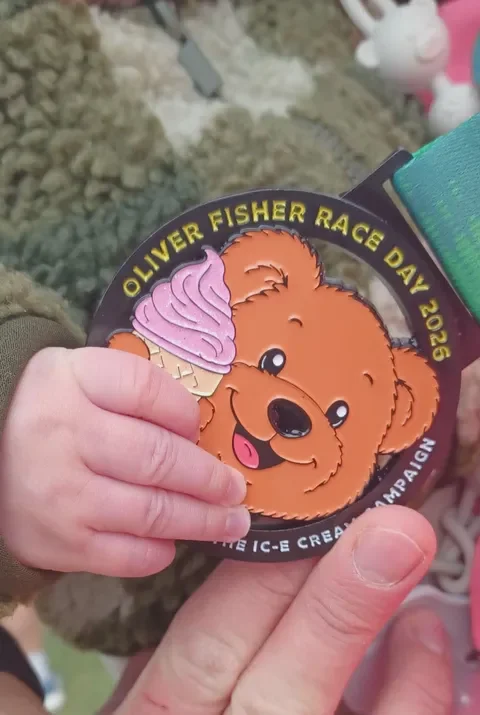 Baby hand holding oliver fisher race day medal with a smiling bear on it