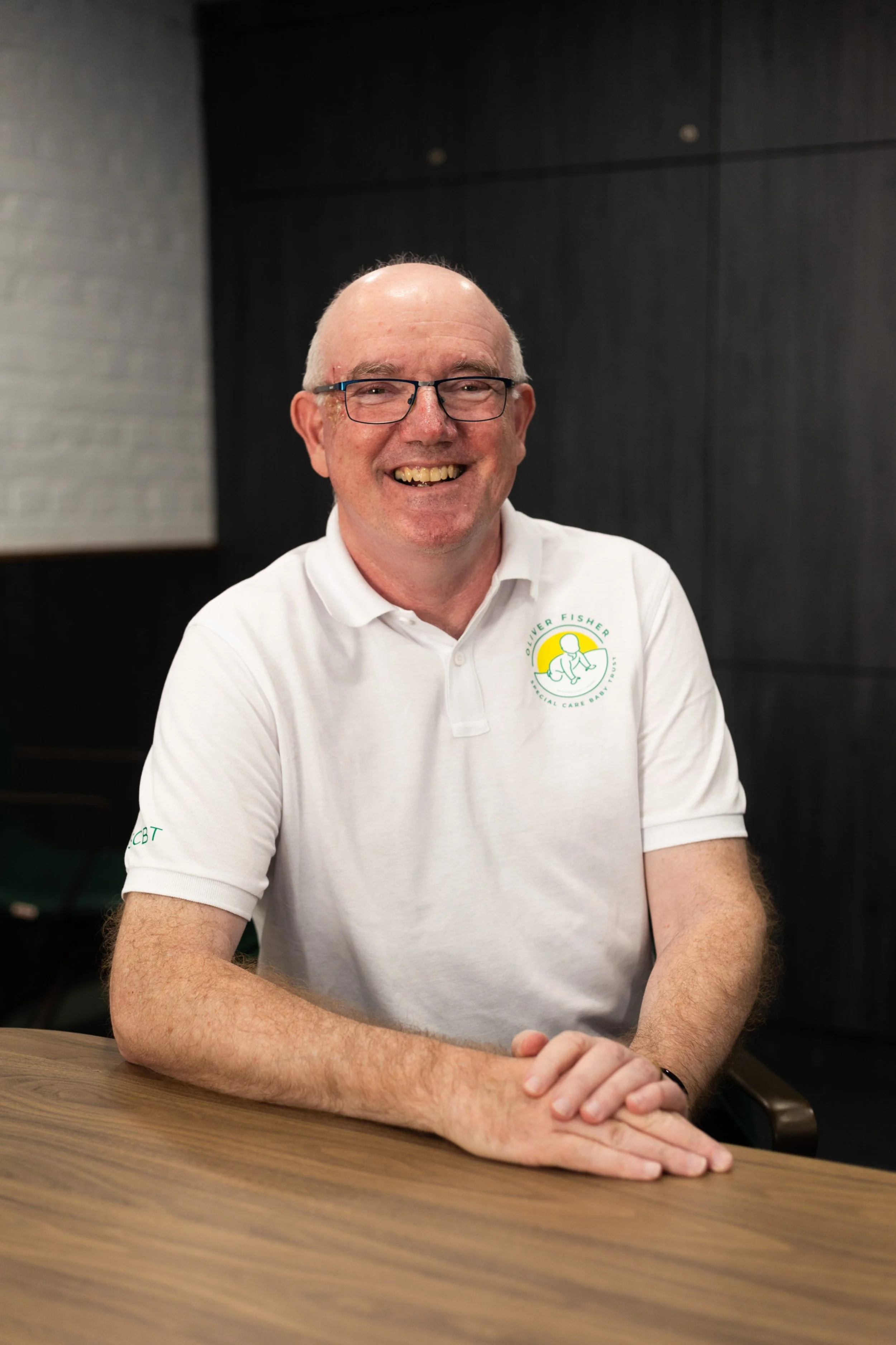 Man smiling wearing white polo shirt with charity logo on plain background