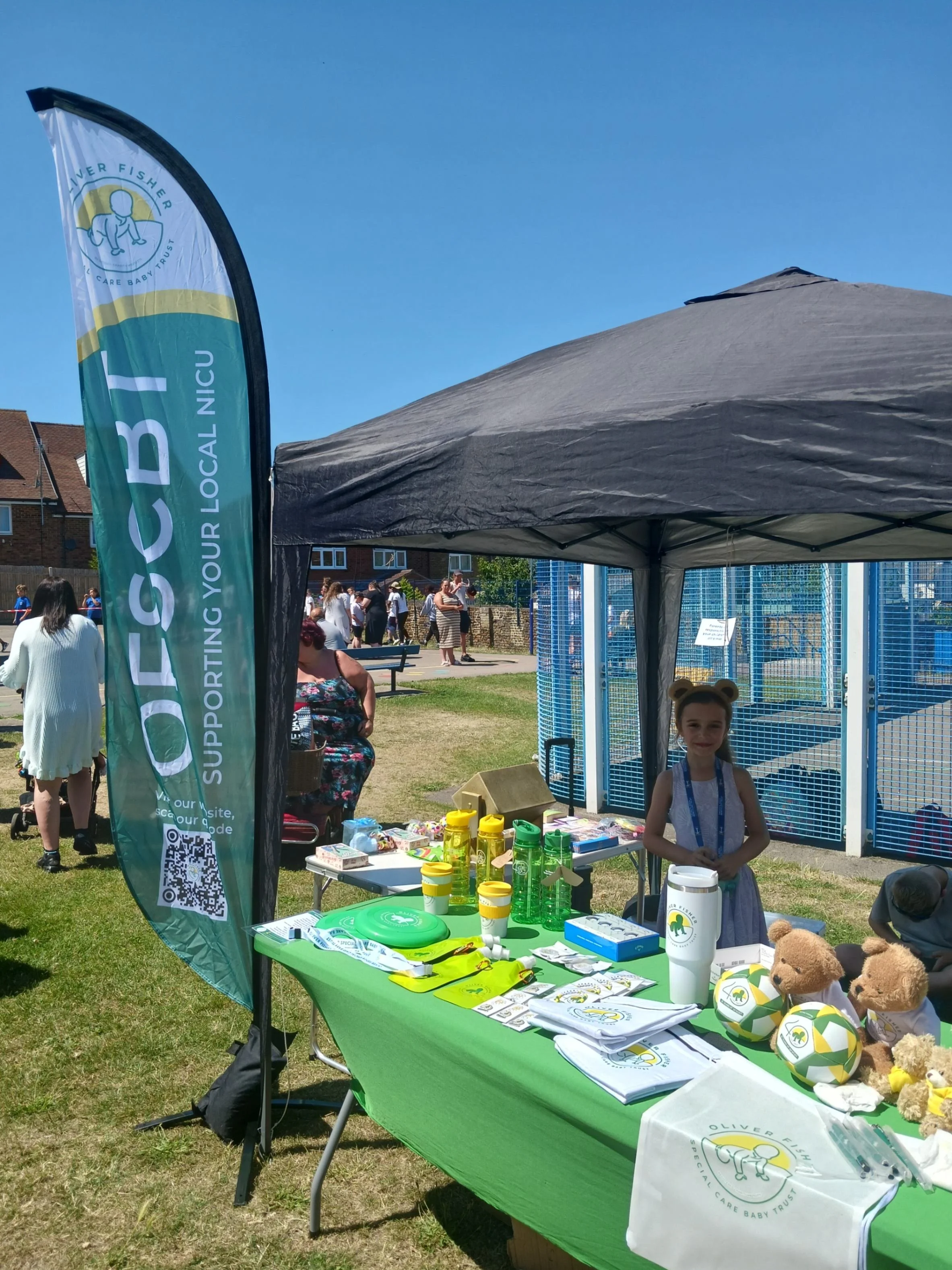 Child standing behind outdoor charity fundraising stall with merchandise and collection items displayed on a table