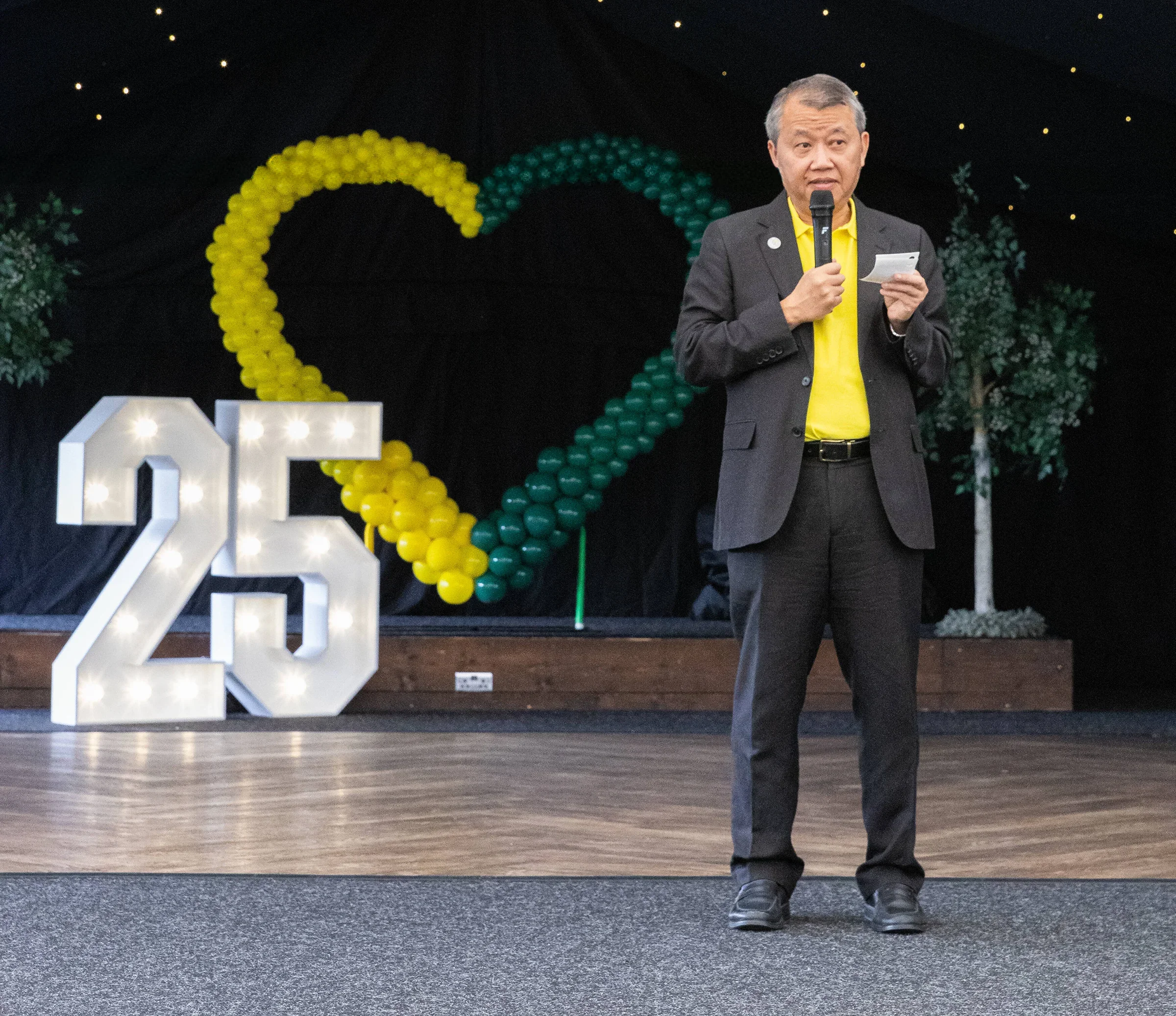 Man speaking into a microphone on stage in front of a large 25 display and balloon decorations