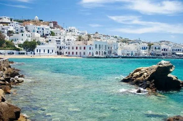 Beautiful coastal scene with white buildings along the shoreline, turquoise water, rocks in the foreground, and a partly cloudy sky.