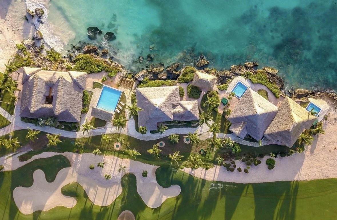 Aerial view of a tropical resort with several thatched-roof buildings, swimming pools, palm trees, and a sandy beach shoreline along turquoise ocean water.