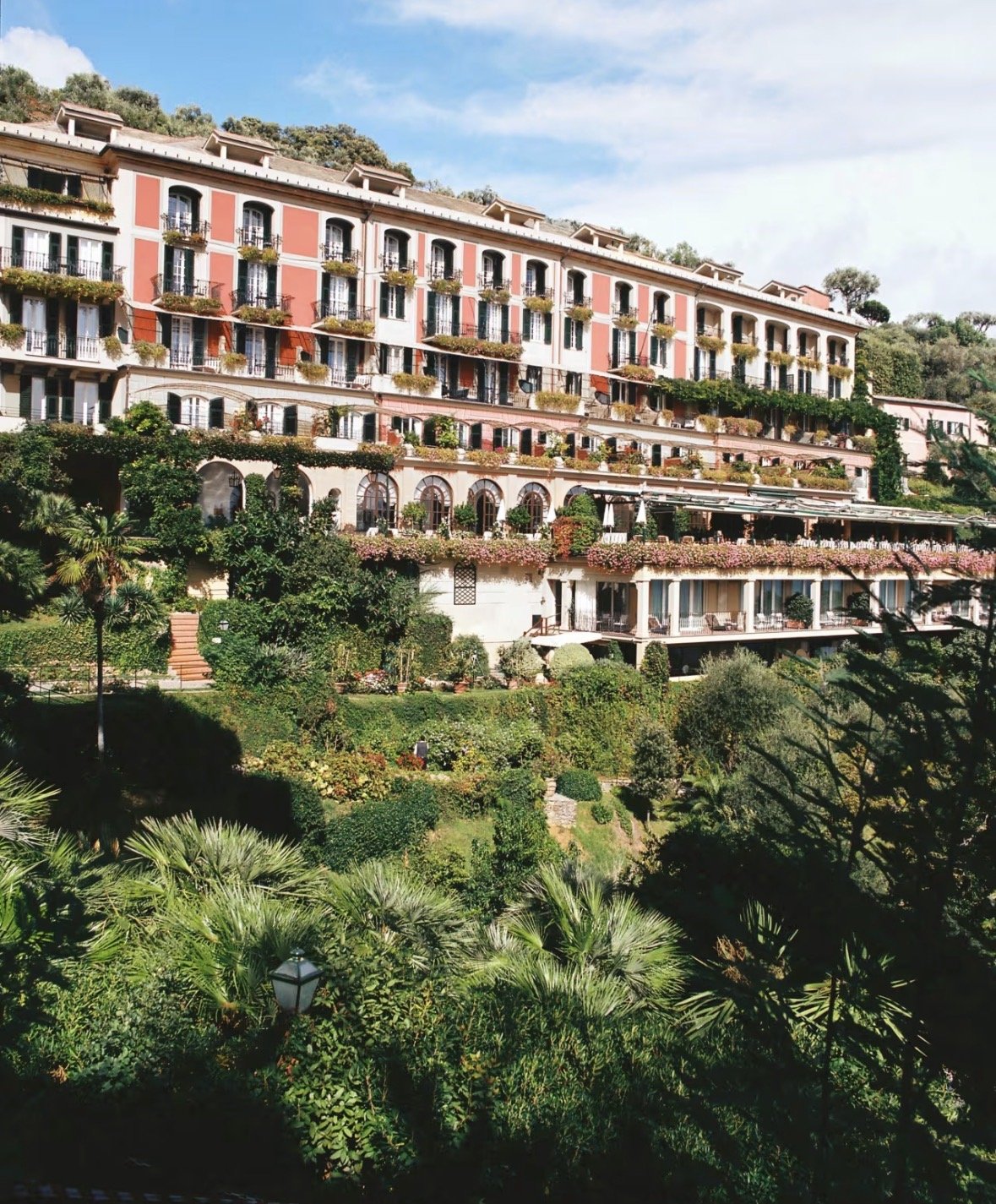 A large multi-story building with pink and white exterior walls, numerous balconies with flower boxes, surrounded by lush greenery and a garden with palm trees, plants, and flowers under a partly cloudy sky.