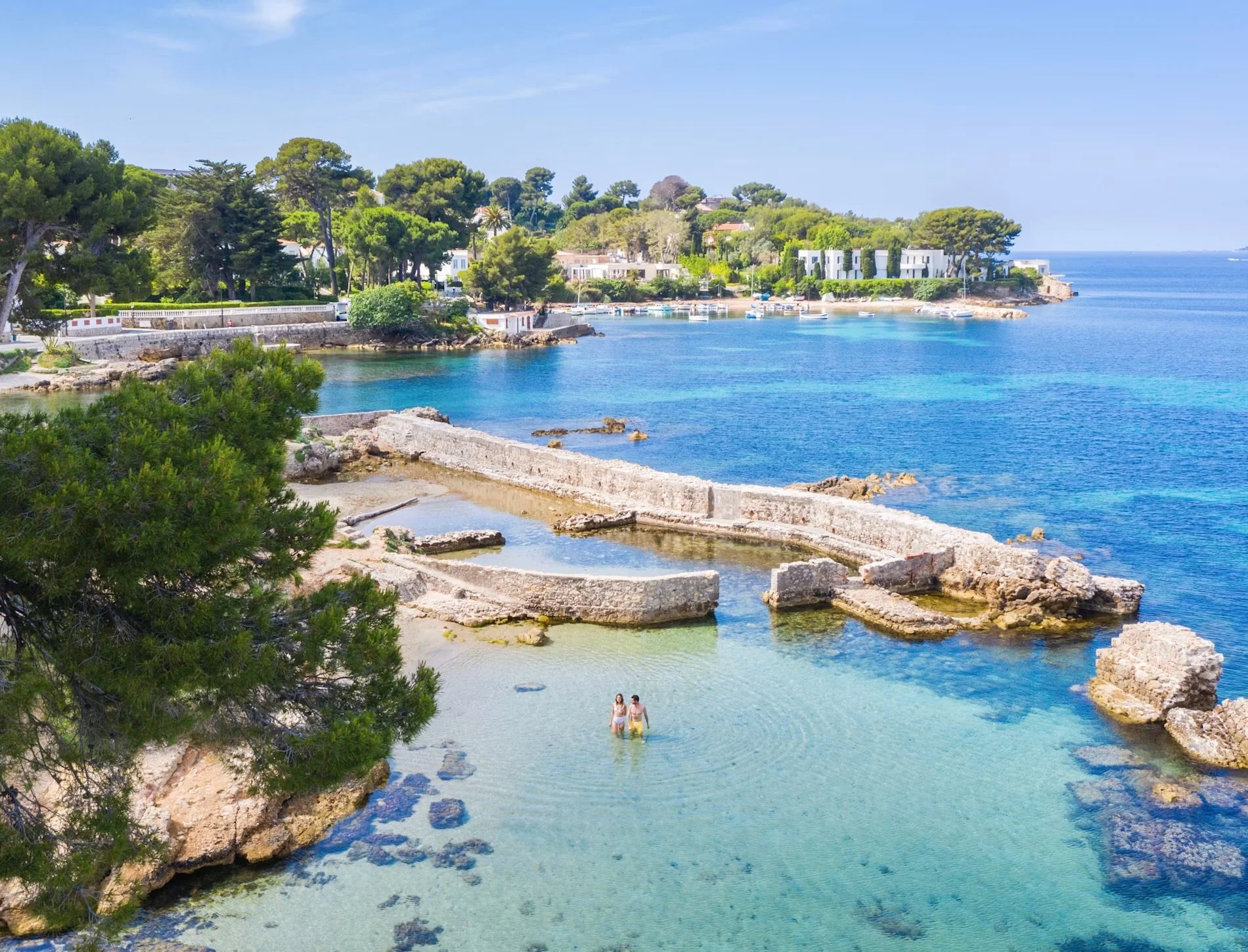 A coastal scene with clear blue water, ancient ruins, trees, and modern houses on a sunny day.