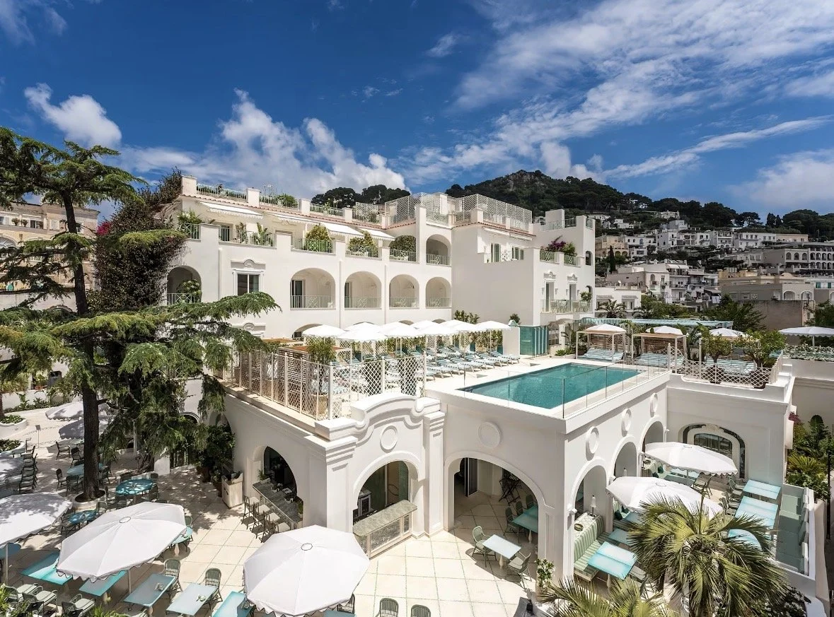 Luxury hotel with a swimming pool, outdoor seating, and white Mediterranean-style architecture against a hillside under a blue sky with clouds.