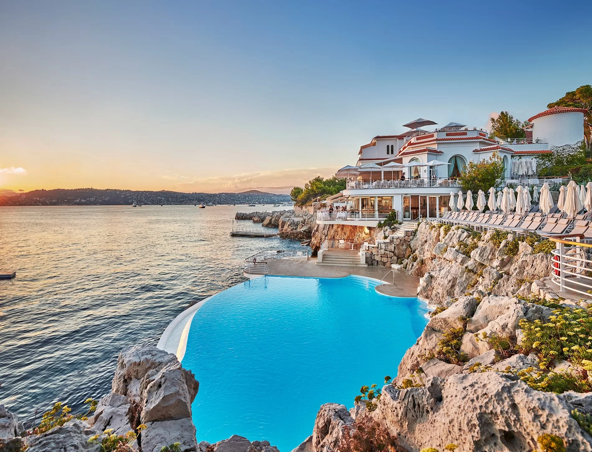 Luxury seaside resort with an infinity pool overlooking the ocean at sunset, featuring a white building with terraces and sun loungers lined up along the rocky shoreline.