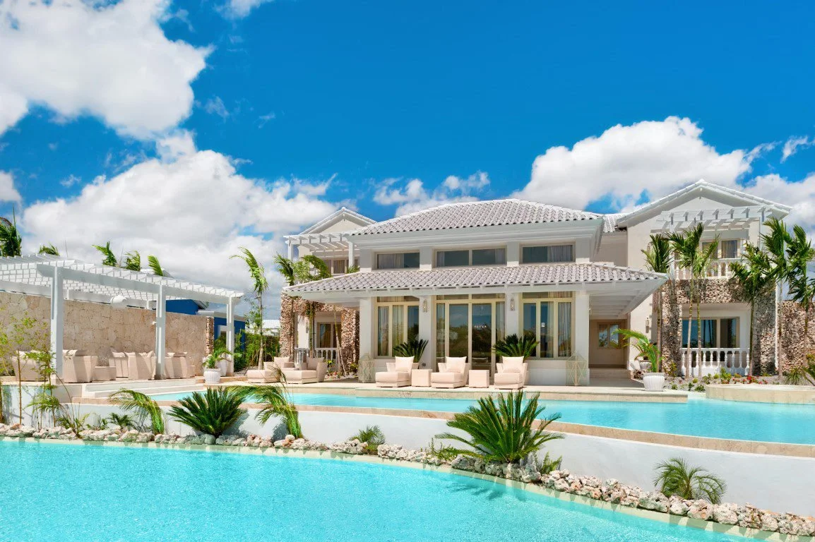 Luxury house with swimming pool, outdoor seating area, palm trees, and blue sky with white clouds.