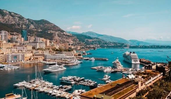 A marina filled with yachts and boats, overlooking a city with buildings and hills in the background, along a body of water.