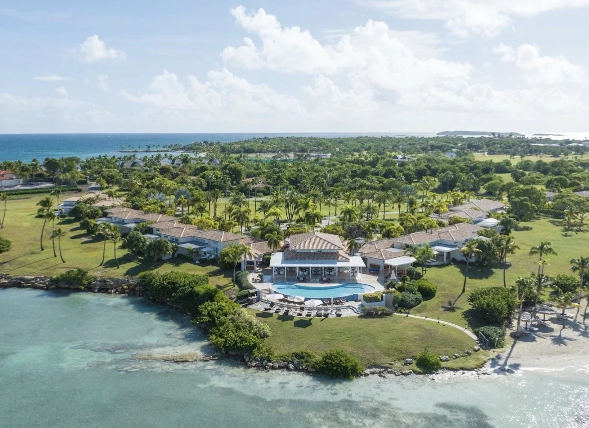 A luxurious beachfront property with a large swimming pool, surrounded by green lawns and tropical palm trees, overlooking the ocean under a partly cloudy sky.