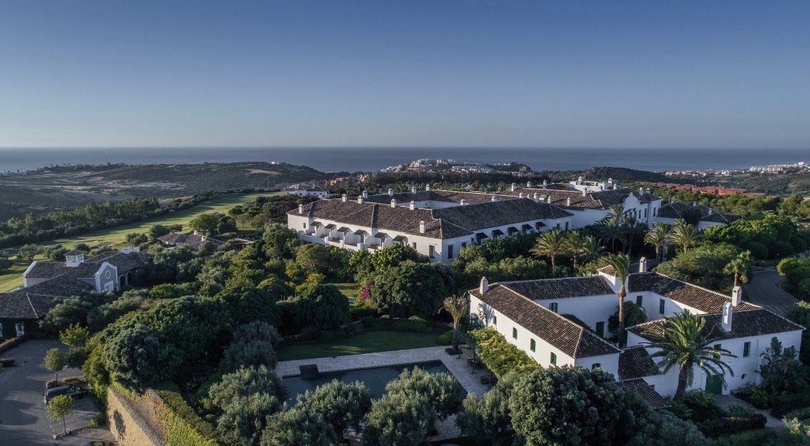 Aerial view of a coastal residential area with white houses with brown tile roofs, lush green trees, and palm trees, overlooking the ocean in the background.