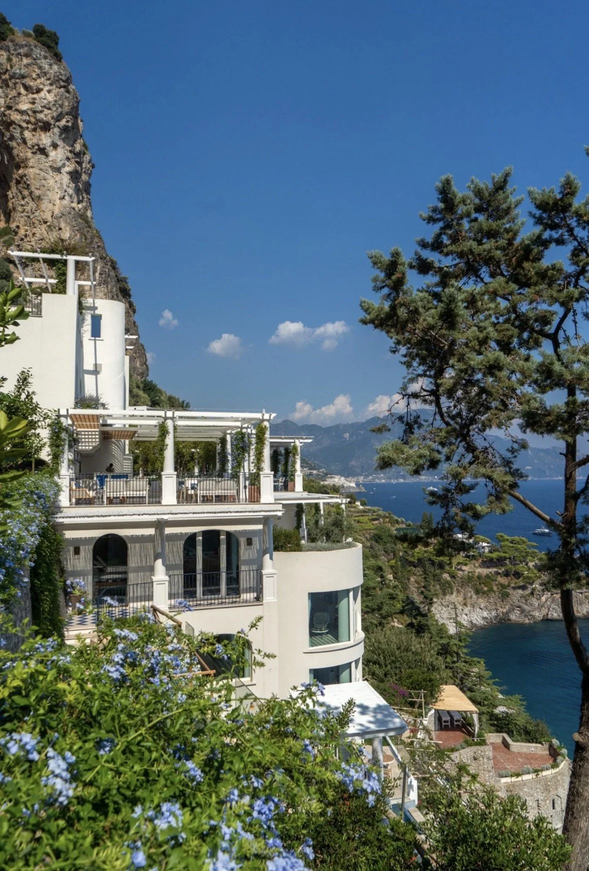 A multi-level white Mediterranean-style house on a hillside overlooking a coastline with blue water and distant mountains, with lush greenery and tall trees nearby.