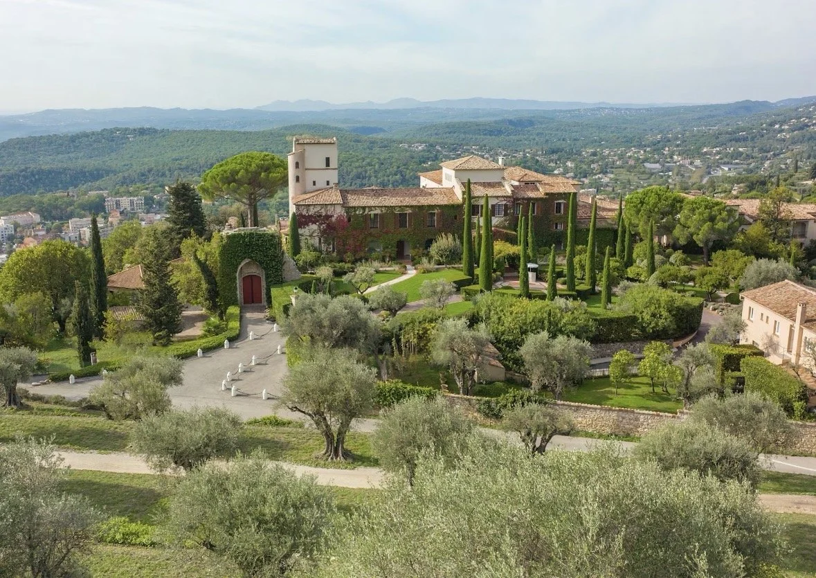 A large estate with many trees, including cypress and olive trees, surrounded by lush green gardens and greenery. The estate features a stone building with a tower and a red door, set in a scenic landscape with rolling hills in the background.