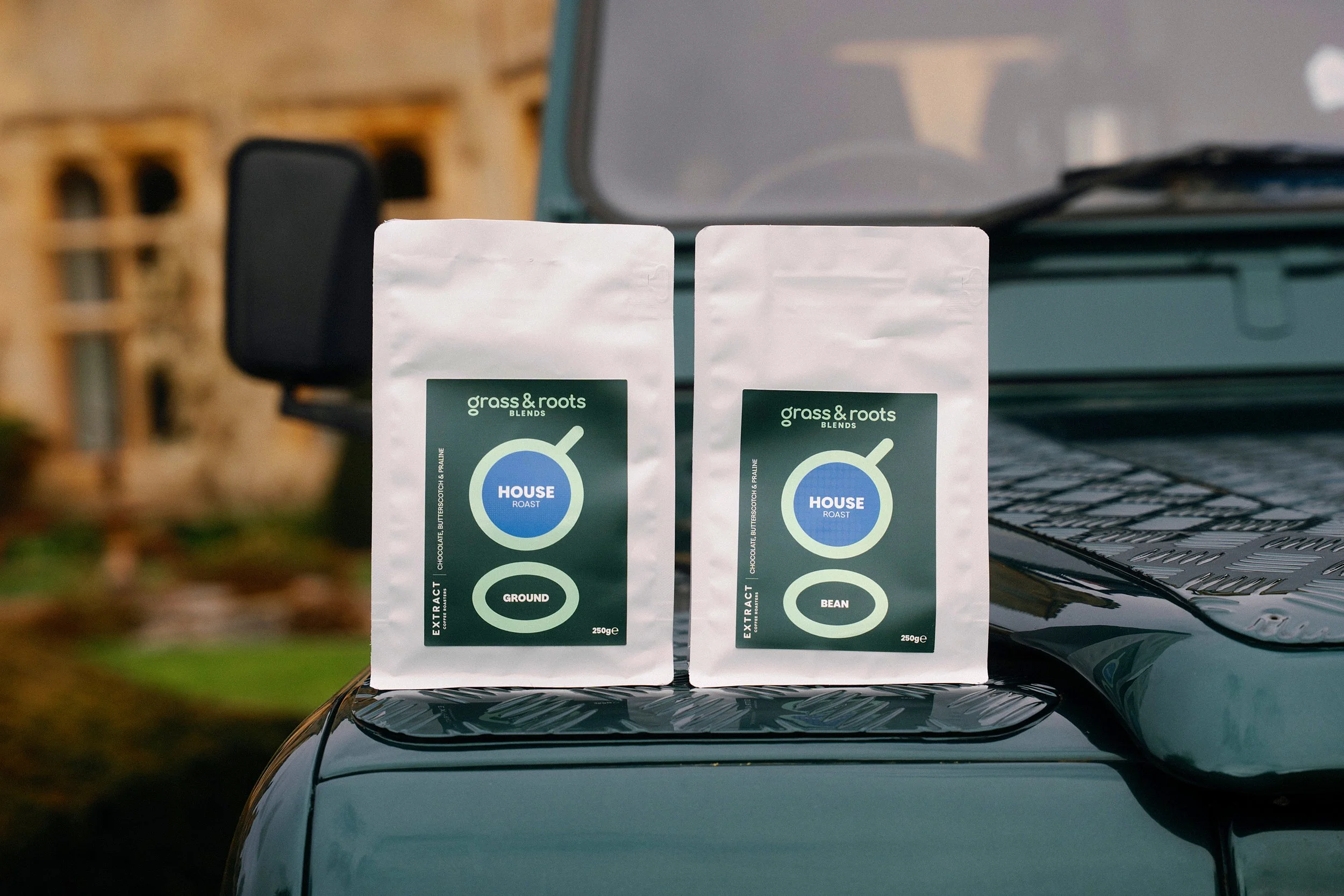 Two bags of grass & roots coffee - one ground and one whole bean - sit on the bonnet of a green Land Rover