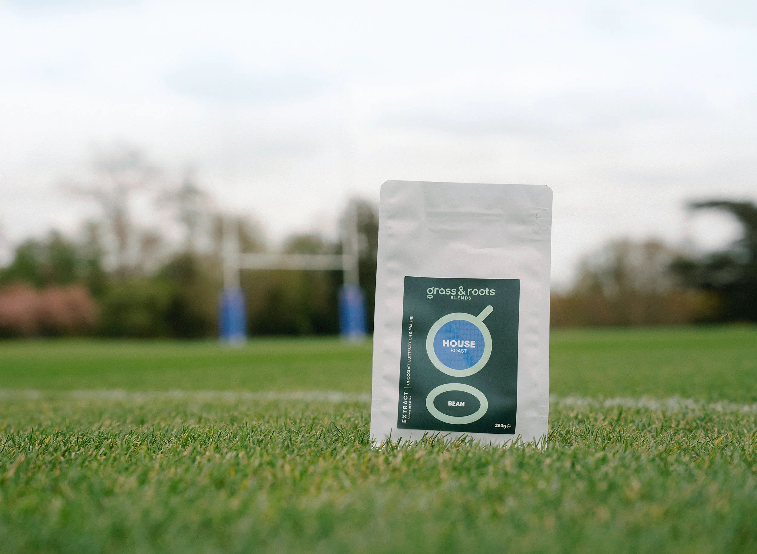 A bag of grass & roots coffee sat on the grass at the edge of a rugby field with some rugby posts in the background