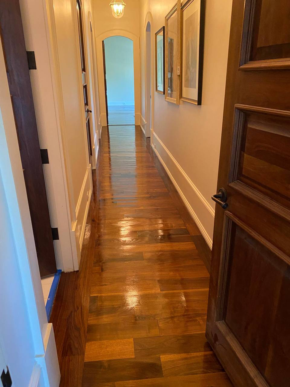A narrow hallway with polished wooden floors, light-colored walls with decorative trim, framed pictures on the right wall, and a ceiling light fixture hanging from an arched ceiling leading to a bright room at the end.